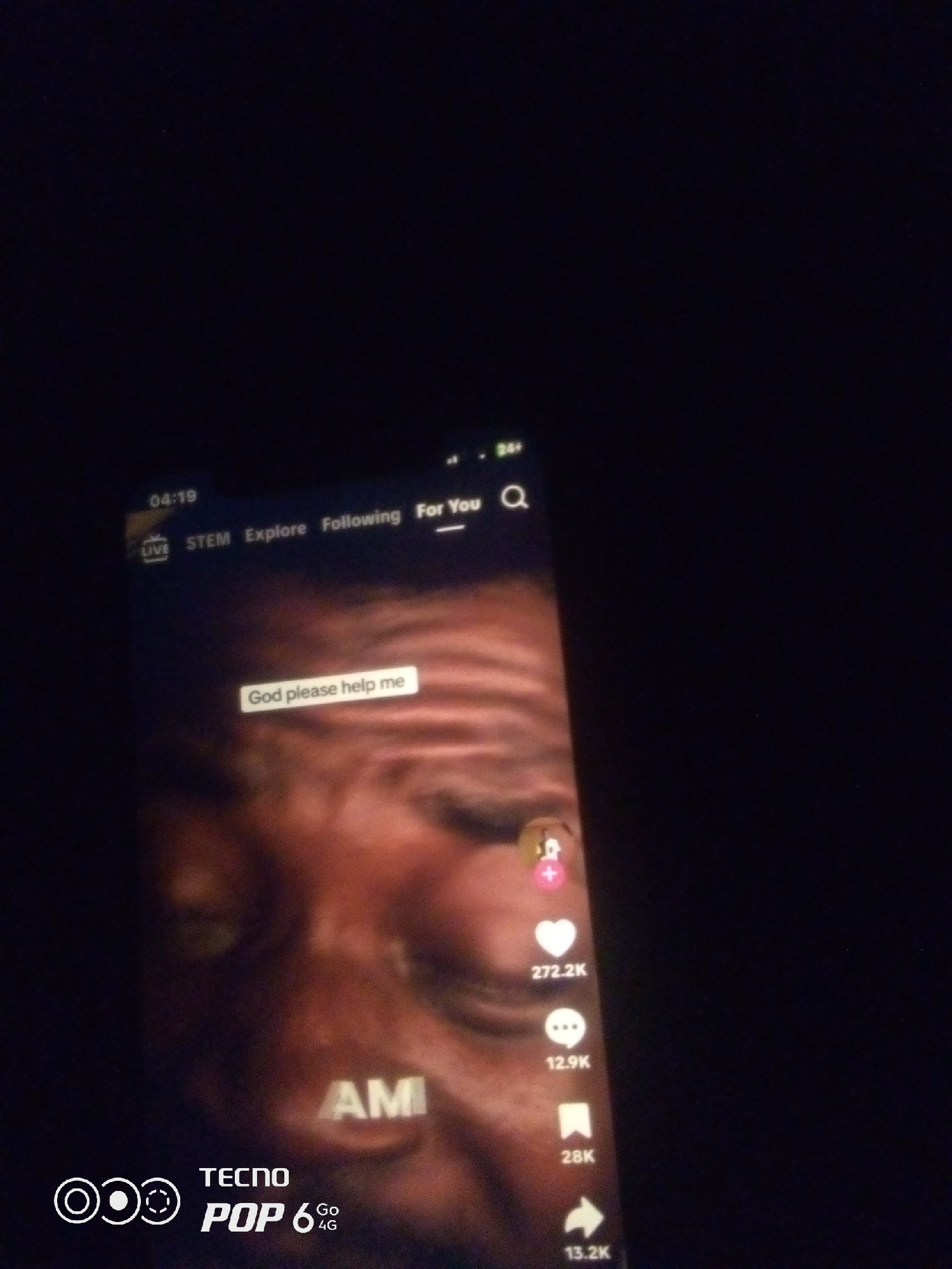 The image shows a close-up of a smartphone screen displaying a social media video. The video appears to be a TikTok or similar platform, with elements like a timestamp "04:19," navigation options "STEM Explore Following For You," and interaction buttons with counts like "272.2K" (likes), "12.9K" (comments), "28K" (shares), and "13.2K" (views). A text overlay on the video reads "God please help me." Another text overlay at the bottom of the screen indicates the phone model as "TECNO POP 6 4G." The background of the video appears to be a close-up of a person's face, with the word "AM" visible on their clothing. The overall lighting is dark, suggesting the photo was taken in low-light conditions. There is no discernible information in the image that places it in Howayi, Nigeria.