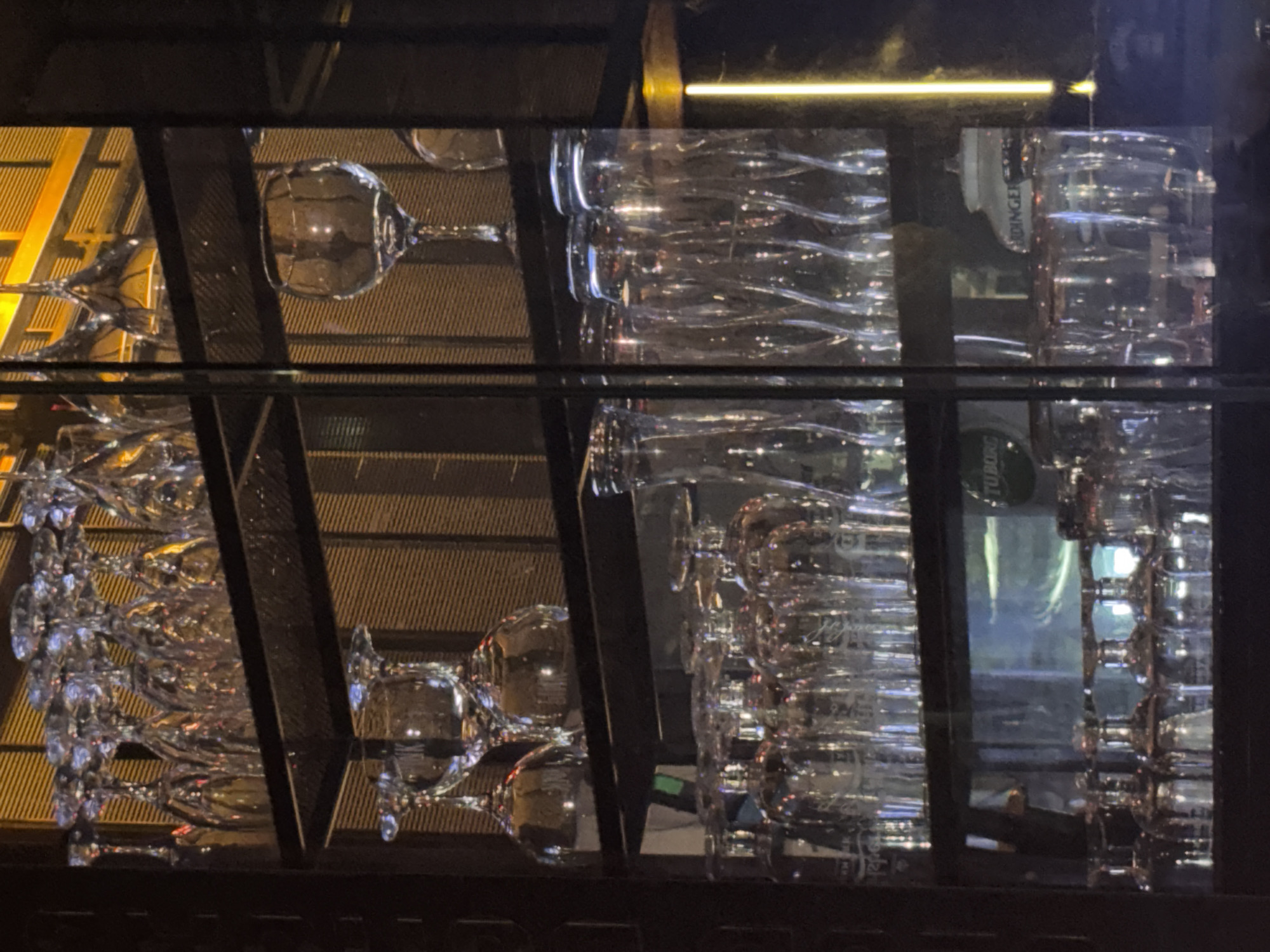 The image shows a shelf of glassware in what appears to be a bar or restaurant. The glassware includes wine glasses, pint glasses, and other drinking vessels. The shelves are made of metal and the background is a textured wall. The lighting is dim, with some warm yellow lights visible. There are reflections in the glass, suggesting it is a display or storage unit. The city of Burgas, Bulgaria is mentioned, but there are no specific visual cues in the image to confirm this location. The image focuses solely on the glassware and its arrangement.