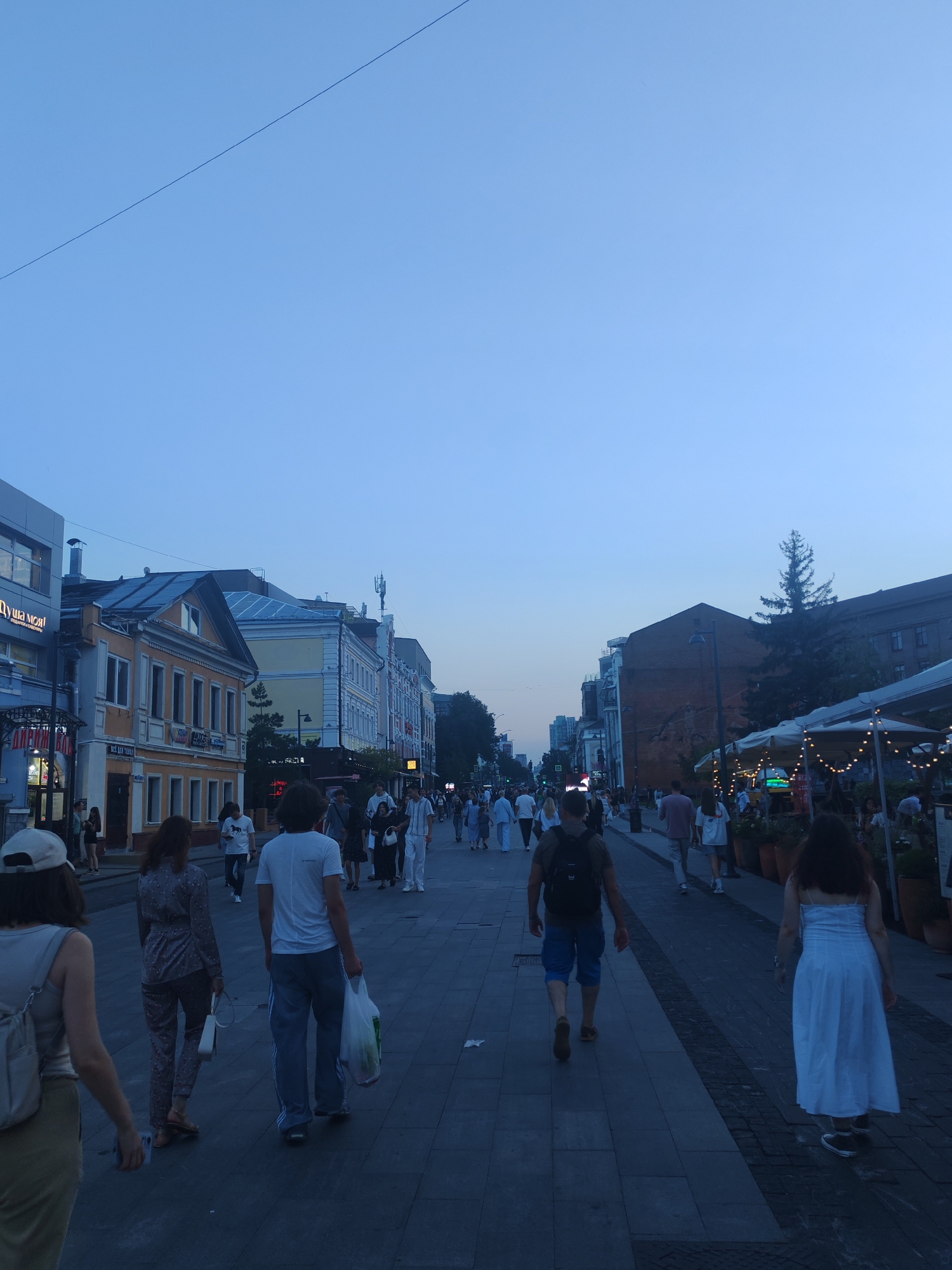 The image captures a street scene in Nizhny Novgorod, Russia, likely during the evening hours, given the twilight sky and artificial lighting visible. The perspective is from ground level, looking down a pedestrianized street lined with buildings on both sides.
On the left, a modern-looking building with signage that includes Cyrillic text is partially visible. Adjacent to it are older, more traditional buildings with yellow and beige facades, featuring multiple windows and what appear to be commercial establishments on the ground floor. Some of these buildings have pitched roofs. Further down the street, more buildings of varying architectural styles can be seen, some appearing more modern.
The right side of the street features buildings, one of which has a brick facade. Several outdoor dining areas with umbrellas and string lights are set up, suggesting restaurants or cafes. A tall, slender evergreen tree stands near the right edge of the frame.
Numerous people are walking along the paved street in both directions, indicating a public space with pedestrian traffic. They are dressed in casual clothing suitable for warm weather. Some individuals are walking alone, while others are in groups. Several people are carrying shopping bags. The ground is paved with cobblestones or similar paving stones, with distinct darker lines indicating sections or seams.
The sky is a clear, pale blue, transitioning to a darker hue towards the top of the frame, indicating the progression of sunset or twilight. A thin, dark line, possibly a power line or cable, stretches diagonally across the upper portion of the sky.
In the foreground, three individuals are prominently featured. On the far left, a person wearing a light-colored top and khaki skirt is walking away from the camera, with a backpack visible. In the middle, a person wearing a patterned pajama-like outfit is walking away from the camera, carrying a white handbag. To the right of the middle person, a man wearing a white t-shirt and blue track pants is walking away from the camera, carrying a transparent plastic bag. Further into the scene, the backs of other pedestrians are visible.
The overall impression is a lively urban environment on a pleasant evening.