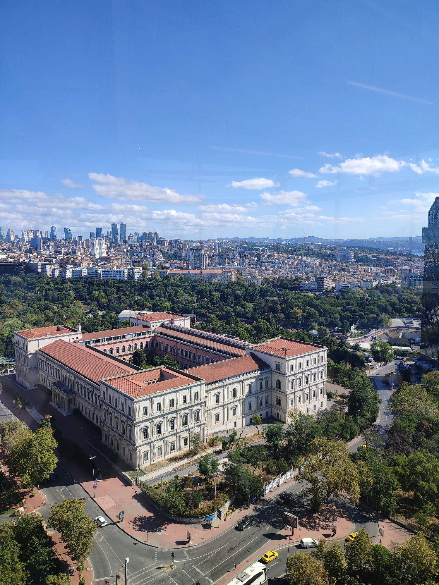 An aerial view captures a sprawling urban landscape under a bright blue sky dotted with fluffy white clouds. In the foreground, a large, pale building with a terracotta tiled roof, featuring numerous arched windows and classical architectural elements, dominates the scene. The building appears to be part of a historical complex, possibly a university or a museum, situated within a lush green park.
Beyond this historic structure, a dense canopy of trees stretches across the middle ground, punctuated by other buildings and a network of roads. Yellow taxis and a white van are visible on the streets, indicating moderate vehicular activity. In the background, a modern cityscape rises, characterized by a cluster of high-rise buildings and skyscrapers that define the city's modern skyline. The distant horizon shows rolling hills and a body of water, suggesting a coastal or riverside location. The scene is bathed in sunlight, casting shadows and highlighting the architectural details of the buildings and the natural textures of the trees. The overall impression is one of a city blending historical charm with contemporary development, set against a backdrop of natural beauty. The location is identified as Beyoğlu, Turkey.
