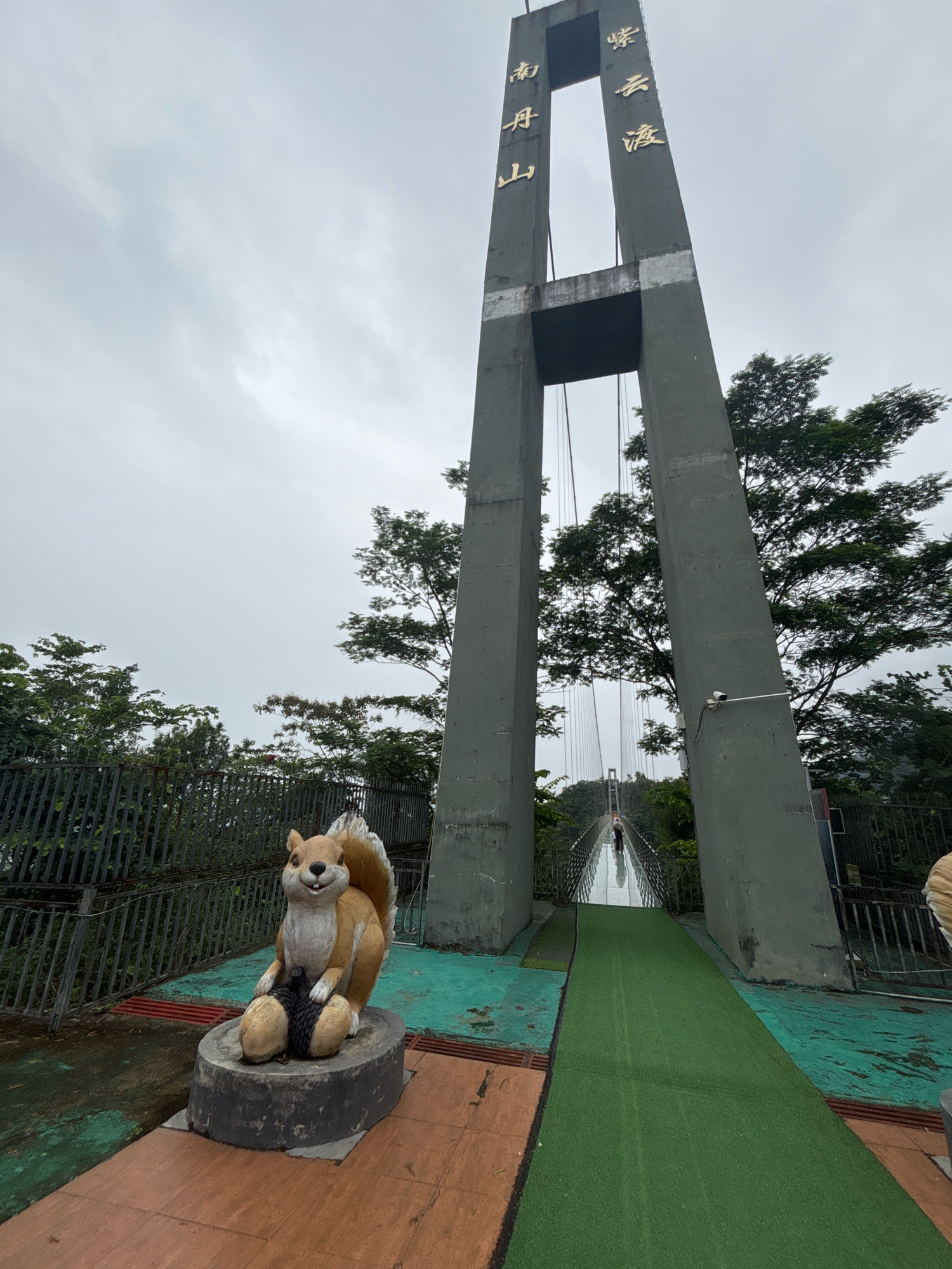 The image depicts a view of a suspension bridge in Foshan, China. The bridge features two large, grey concrete pylons that support the structure. Golden Chinese characters are inscribed on the pylons. In the foreground, on the left side of the frame, is a statue of a squirrel sitting on a base and holding two acorns. The bridge walkway is covered in green turf leading towards a person walking in the distance. The sky is overcast and grey, and trees are visible on either side of the bridge. A metal fence borders the area.