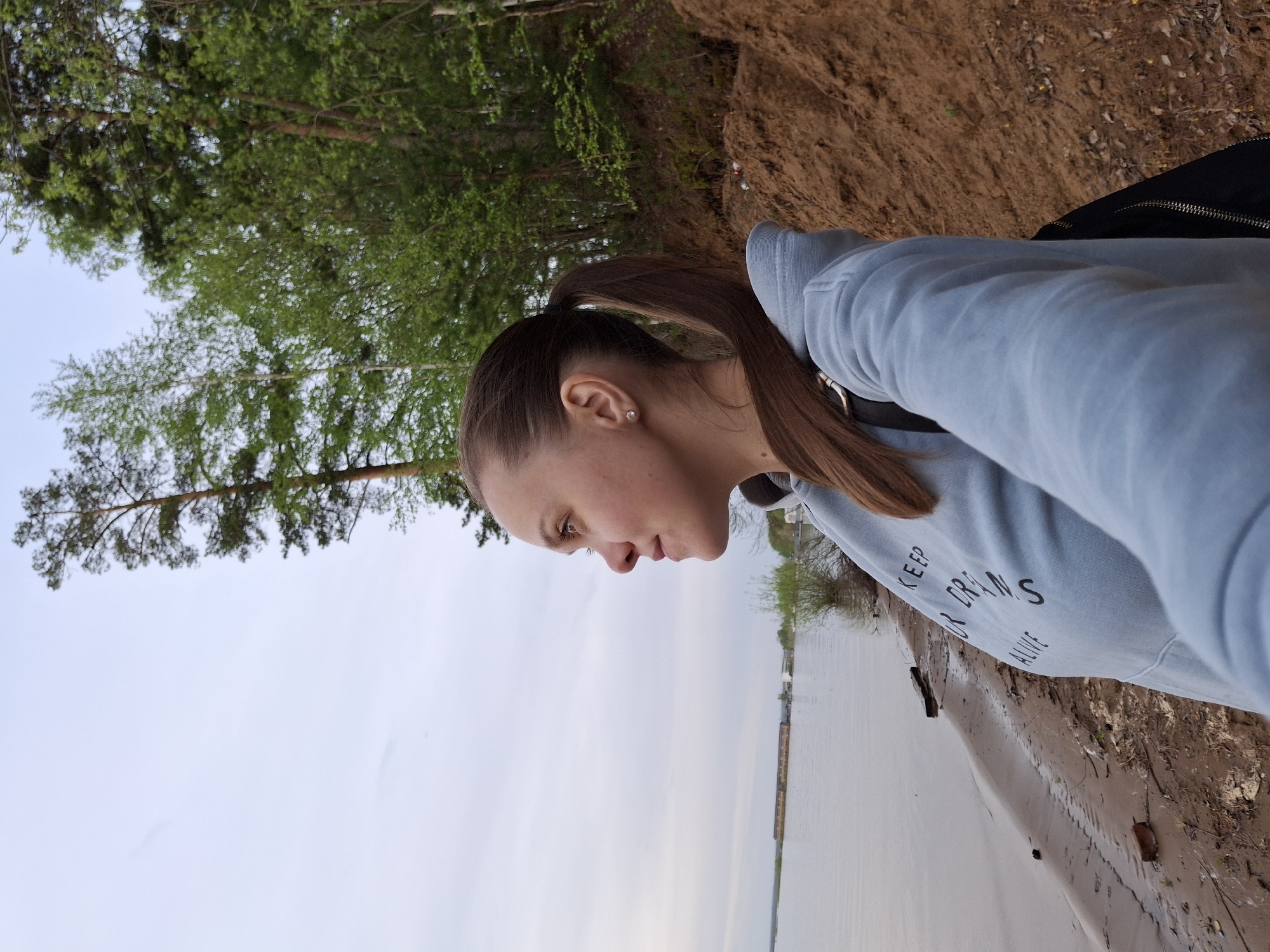 A woman with light skin and brown hair pulled back into a ponytail is standing near the edge of a body of water. She is wearing a light blue hooded sweatshirt with white lettering that reads "KEEP YOUR DREAMS ALIVE". The background consists of trees and a bank of sandy or earthen material. In the distance, buildings can be seen across the water. The scene appears to be in Chaikovsky, Russia.