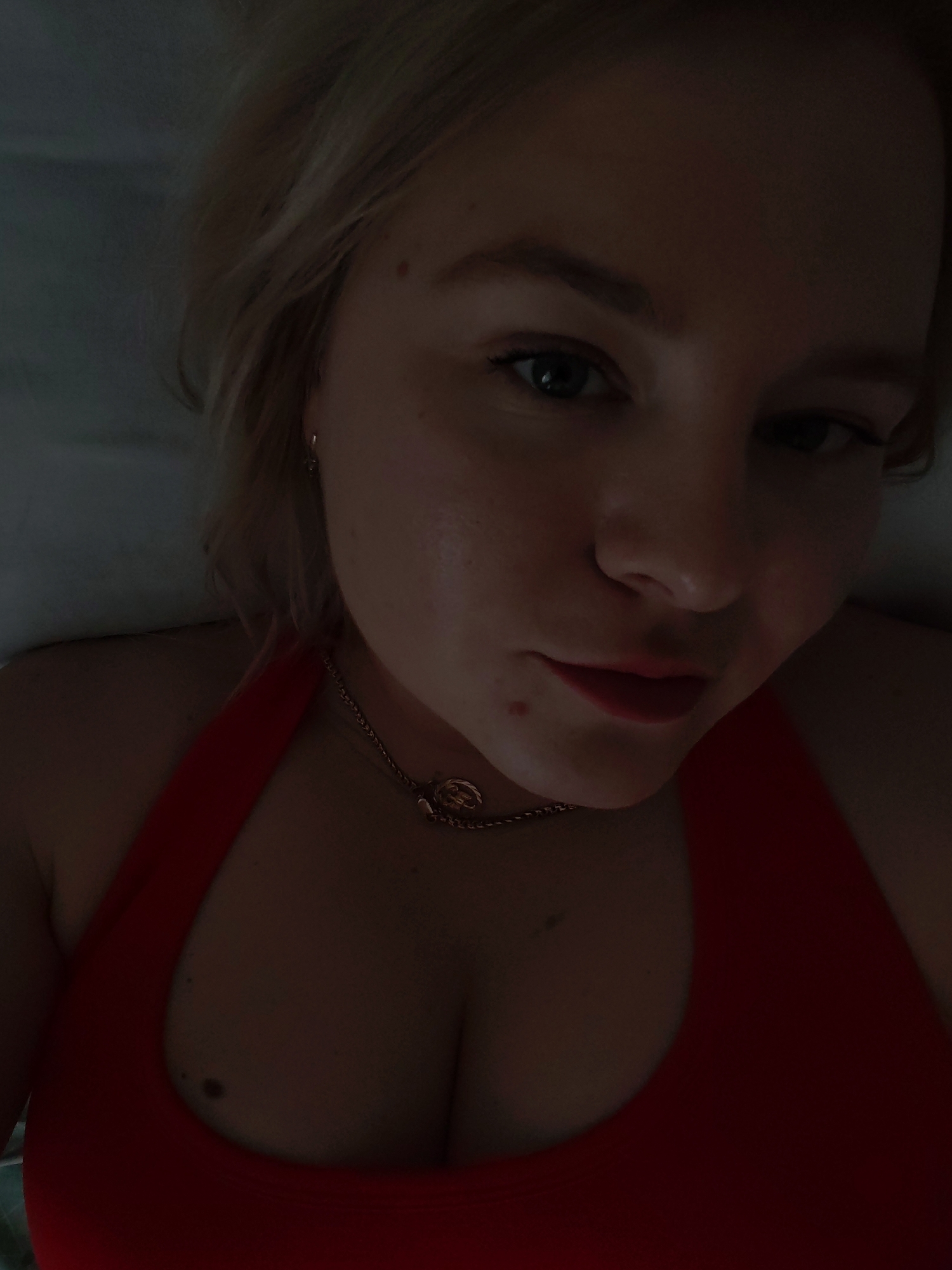 A close-up selfie of a fair-skinned woman with blonde hair. She is wearing a red halter-neck top and a gold necklace. The background is out of focus, appearing dark. The location of the city of Izhevsk, Russia is not evident from the image.