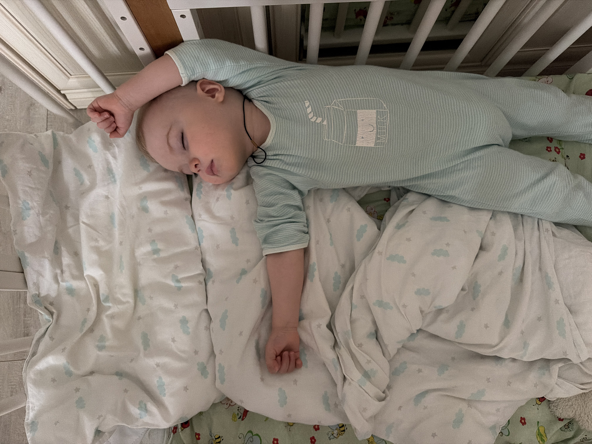 A baby is asleep in a crib. The baby is wearing a light blue striped onesie with a milk carton graphic. The crib has white railings and decorative panels. The bedding includes a white pillowcase with light blue cloud and star designs, and a patterned sheet beneath with floral and bee motifs. A thin black cord is around the baby's neck. The location is unknown and cannot be determined from the image. There is no way to ascertain that the picture was taken in Votkinsk, Russia.