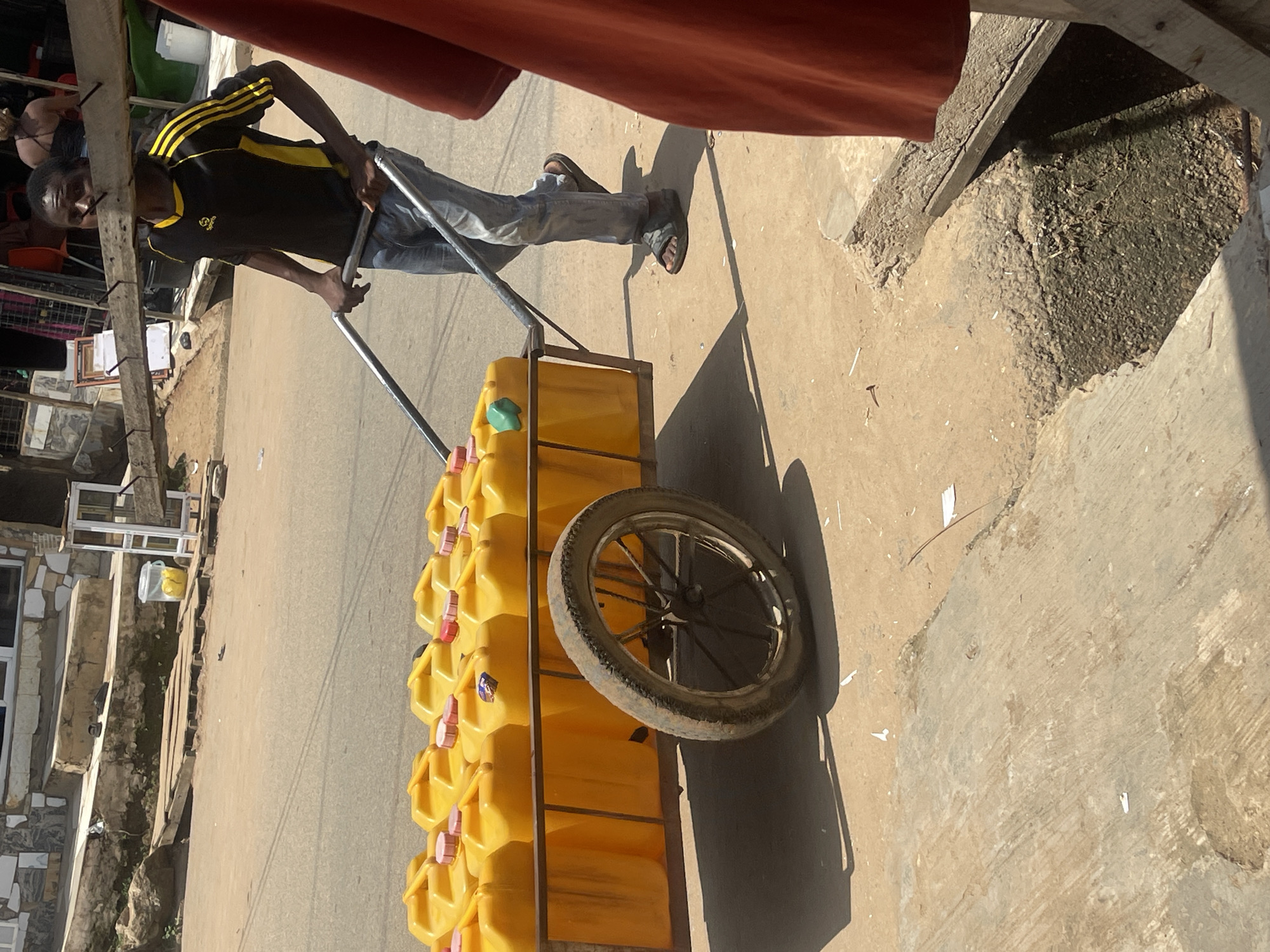 A young Black male is pushing a cart loaded with yellow jerrycans on a paved street. The cart has a single wheel and two metal handles. The jerrycans are stacked two high, with red lids on most of them. The man is wearing a black t-shirt with yellow stripes on the sleeves and dark blue jeans. He is also wearing dark sandals. In the background, there are buildings and other people. The scene appears to be in a market or commercial area. The location is identified as Abuja, Nigeria.