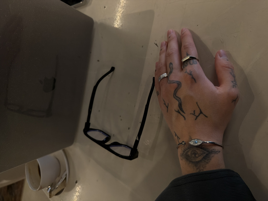 The scene depicts a close-up, overhead view of a person's left hand and forearm resting on a light-colored table in Yerevan, Armenia. The visible hand and forearm exhibit multiple tattoos, including a large eye design on the inner forearm, a serpentine line across the back of the hand, and several abstract, linear markings on the hand and fingers. Two silver-colored rings are worn: a textured band on the ring finger and a smooth, geometric band on the pinky finger. A silver cuff bracelet featuring an oval, iridescent "evil eye" charm is present on the wrist. On the table, adjacent to the hand, are a pair of black-framed eyeglasses with light blue lenses. Behind the eyeglasses, a silver-grey laptop is positioned, with a faint Apple logo visible as a reflection on its closed lid. In the upper left corner of the frame, a white ceramic coffee cup containing dark liquid sits on a white saucer, accompanied by a silver spoon and a white paper packet.