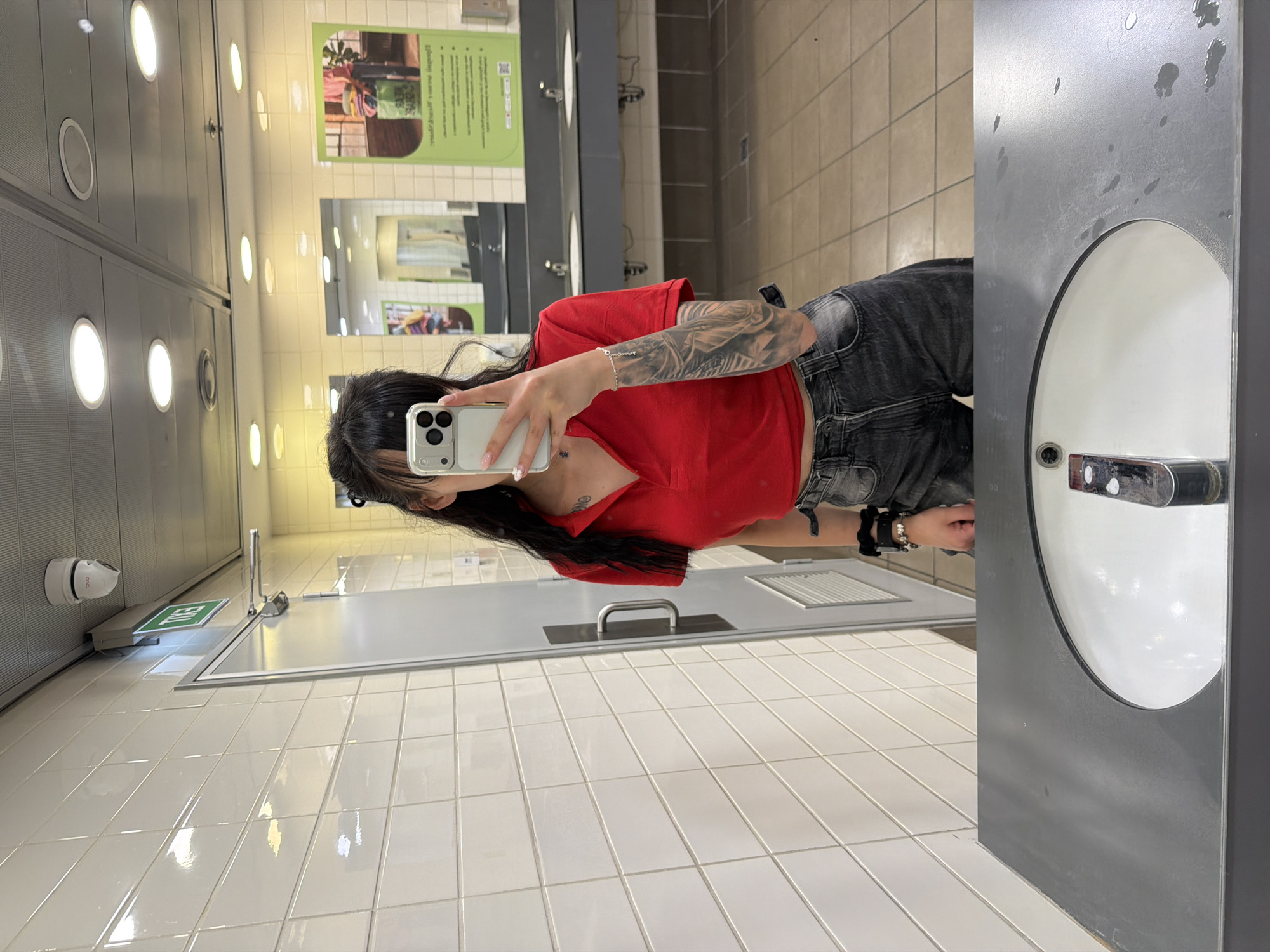 The image depicts a young woman taking a mirror selfie in a public restroom. She is wearing a red polo shirt and black denim shorts. Her left arm is covered in tattoos. The restroom has white tiled walls and beige tiled floors. There are multiple sinks visible in the background, along with a large mirror. A security camera is mounted on the ceiling in the upper left corner. An exit sign is visible above a door. The overall impression is of a typical public facility. The city of Plovdiv, Bulgaria, is not depicted in the image, nor are there any elements that would suggest this specific location.