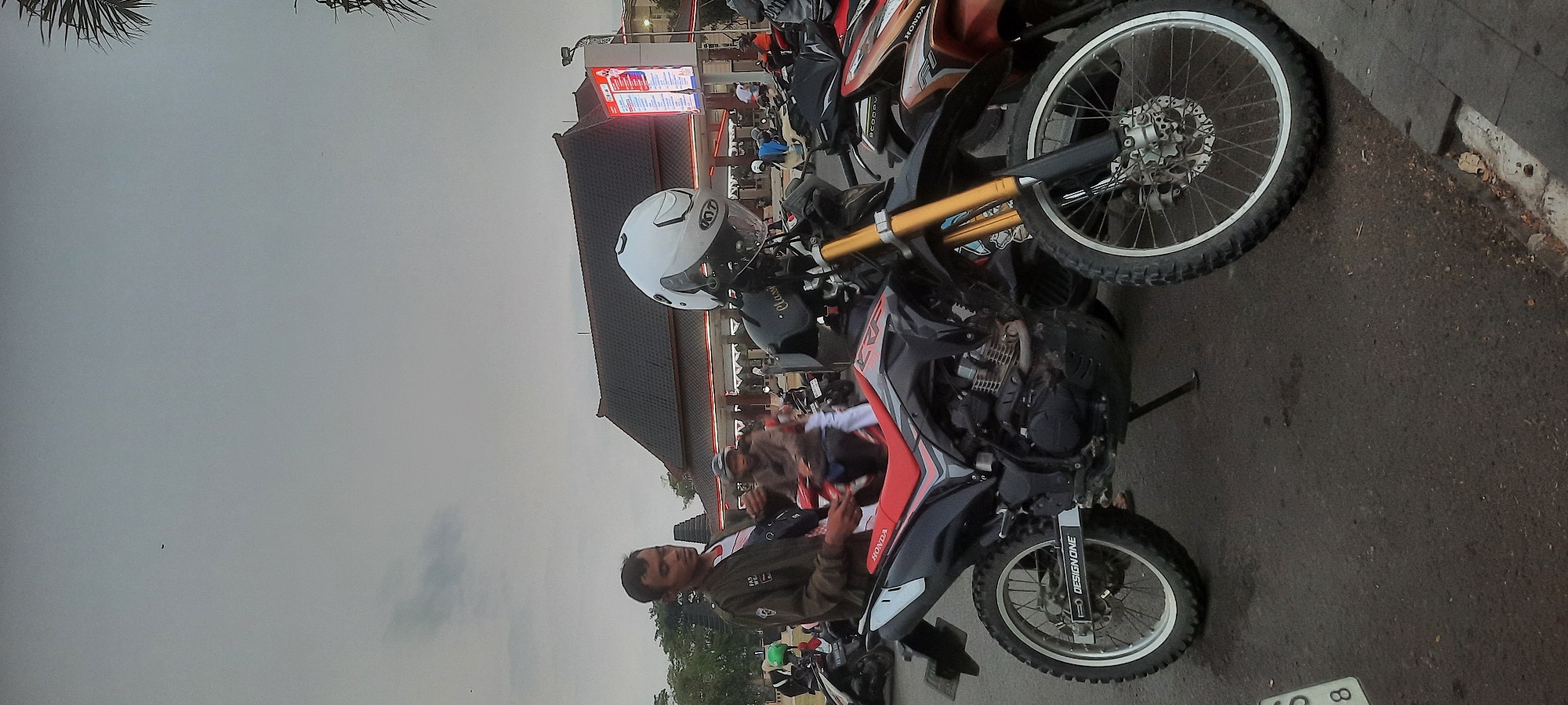 In Kanigoro, Indonesia, under a cloudy sky, a man stands next to a dirt bike parked on a paved surface. The motorcycle is black and red, with gold front suspension and knobby tires. A white helmet with a "KYT" logo rests on the handlebars. In the background, a traditional building with a dark tiled roof is visible, along with other parked motorcycles and people. The scene appears to be a public area, possibly a parking lot or a gathering spot.