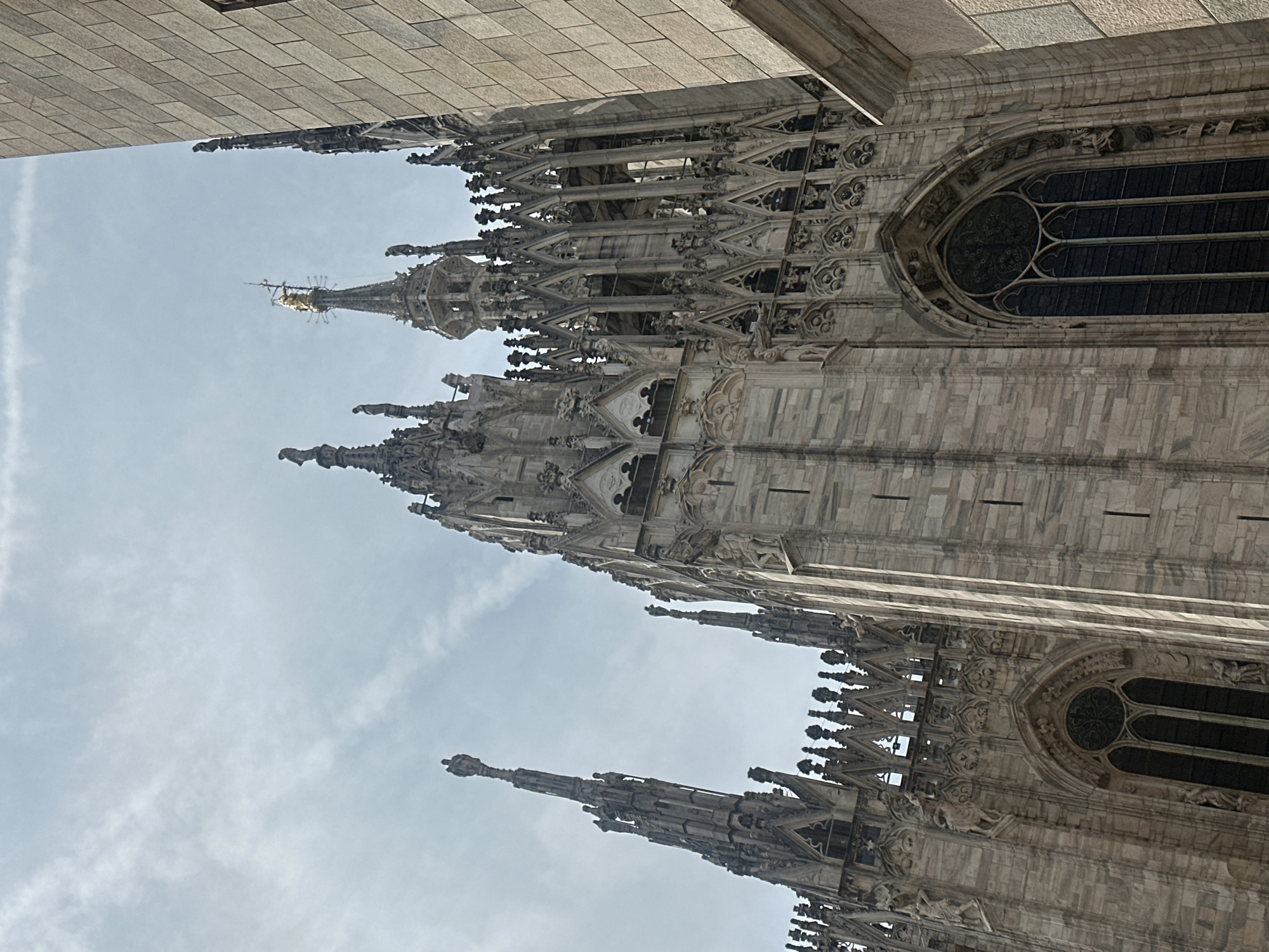 The image shows a close-up view of the Duomo di Milano, a cathedral in Milan, Italy. The architecture is Gothic, characterized by intricate spires, pointed arches, and ornate stonework. The facade is made of marble, with statues and decorative elements adorning the exterior. The sky is partly cloudy, with a pale blue hue. The lighting suggests it is daytime, likely late morning or early afternoon, casting soft shadows on the building. The perspective is from below, looking upwards, emphasizing the scale and grandeur of the cathedral. The right side of the image shows a section of the cathedral's side wall, with a distinctly different, more modern stone facade, also made of large, light-colored rectangular blocks. The contrast in the materials suggests a later addition or renovation to the original structure.