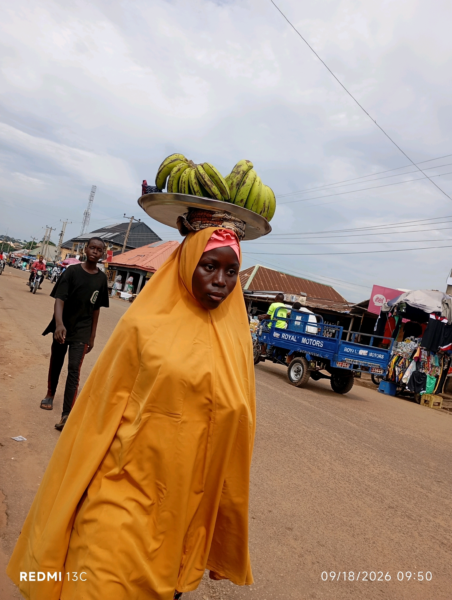 In the city of Shendam, Nigeria, a young woman wearing a yellow hijab and a flowing yellow garment walks down a dirt road. She carries a tray of green bananas balanced on her head. To her left, a young boy in a black t-shirt and dark pants walks in the same direction. In the background, a blue truck with "ROYAL MOTORS" written on its side is parked on the road. Several market stalls with various goods are visible, and other people and motorbikes are present in the distance. The sky is overcast. The timestamp 09/18/2026 09:50 is visible in the lower right corner.