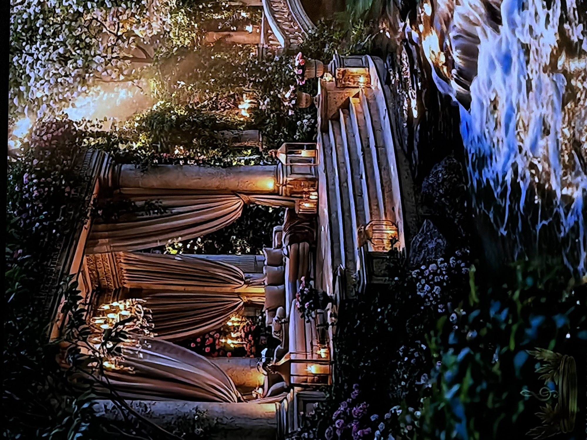 This image depicts an ornate outdoor garden or event space illuminated at night, viewed as if rotated 90 degrees clockwise. The scene is rich with classical architectural elements, abundant foliage, and decorative lighting.
On the left side of the upright image, a grand structure features tall, fluted columns supporting intricately draped, striped fabrics, possibly part of a pavilion or archway. Within and around these drapes, numerous warm, glowing lights, likely candles or lanterns, are visible, alongside clusters of white and pink flowers. Below, a series of light-colored cushions suggests a seating area.
Towards the center, a broad set of stairs or a terraced pathway ascends, flanked by similar architectural columns and decorative lanterns that cast a soft, inviting glow. This path is bordered by lush greenery and flowering plants, primarily with small white and pink blossoms.
On the right side, a striking waterfall or large fountain cascades downwards, its water dramatically illuminated with a luminous blue-white light that contrasts sharply with the warm tones elsewhere. The immediate foreground and background areas are densely populated with dark green shrubs and small, light-colored flowers.
The setting evokes an atmosphere of elegance, romance, and festivity, typical of a lavish outdoor event or a beautifully maintained garden at night. There are no people visible, and no discernible text. The location context provided is Sofiivska Borshchahivka, Ukraine.