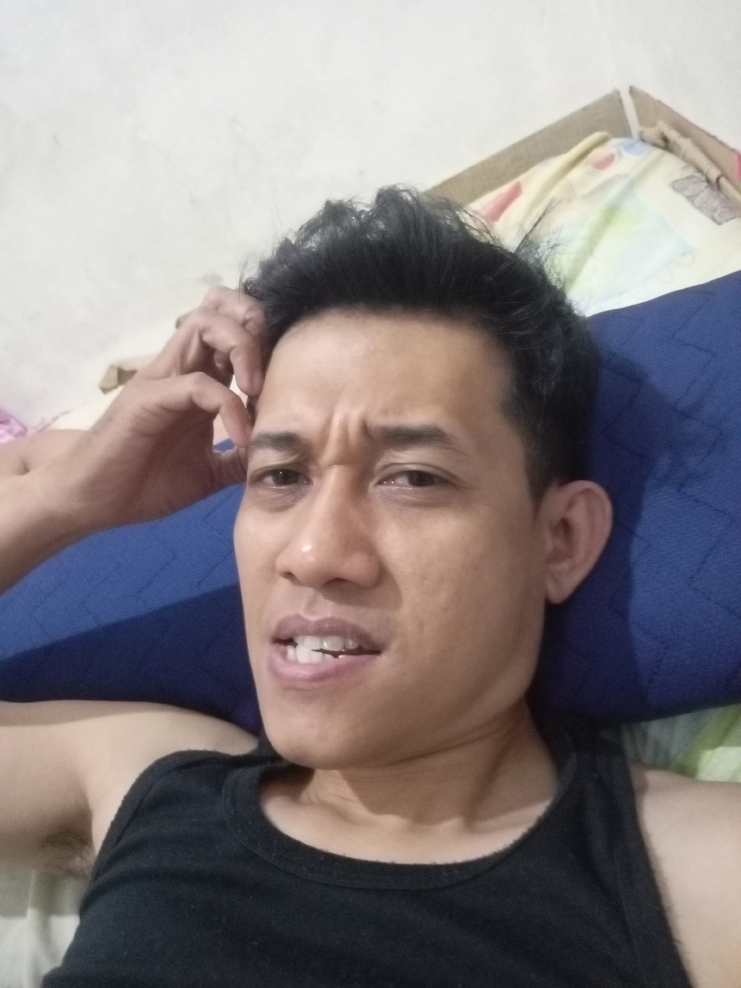 The image is a selfie taken indoors, likely in a bedroom, by a man who appears to be lying down. He has dark, short, messy hair, and is wearing a black tank top. His left hand is raised to his forehead, and his facial expression suggests confusion or contemplation, with furrowed brows and a slightly open mouth revealing his teeth. The background shows a blue pillow, a patterned bedsheet, and a white wall. The quality of the image is slightly grainy, common for personal photographs taken with a mobile device. While the exact location within Batam, Indonesia cannot be determined from the visual information alone, the scene depicts a private, domestic setting.