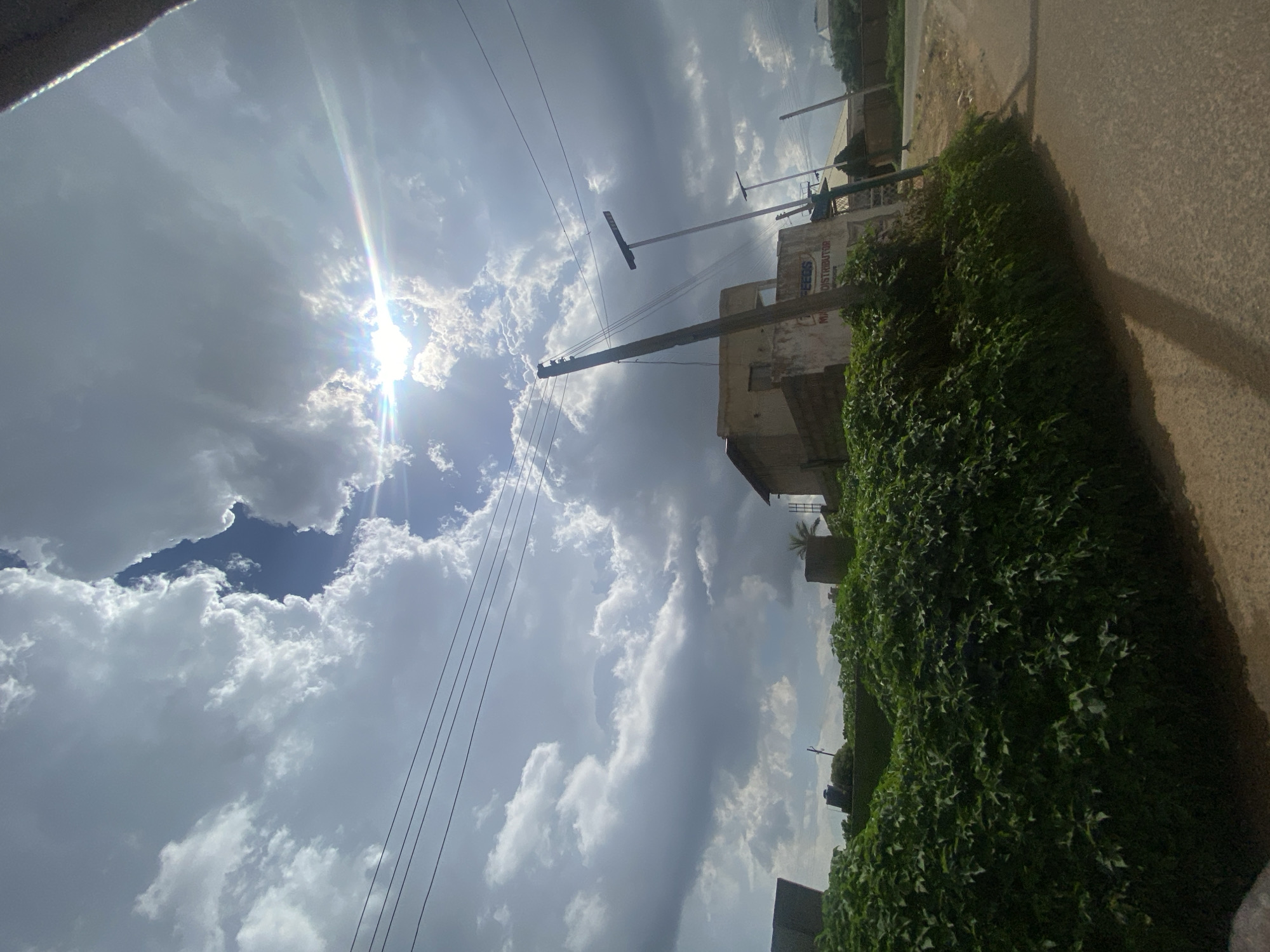 The image depicts a street scene in Jos, Nigeria, under a cloudy sky with the sun peeking through. The foreground is dominated by a dense, green hedge of foliage that borders a dusty, paved road. To the right of the road, the terrain slopes slightly upwards, with another paved area visible further down. In the middle ground, a building with light-colored walls stands partially obscured by the vegetation, displaying signage that reads "FEEDS MA DISTRIBUTOR." Several utility poles, one of which is leaning, are positioned near the building, with overhead wires crisscrossing the frame. Three solar-powered streetlights with rectangular panels are also visible, along with another utility pole further in the background. The sky is dramatic, with large, billowing clouds creating a dynamic contrast with the bright sunlight, which casts a lens flare across the image. The overall impression is one of an urban or semi-urban environment in Nigeria, likely during the daytime, characterized by its vegetation, infrastructure, and weather conditions.
