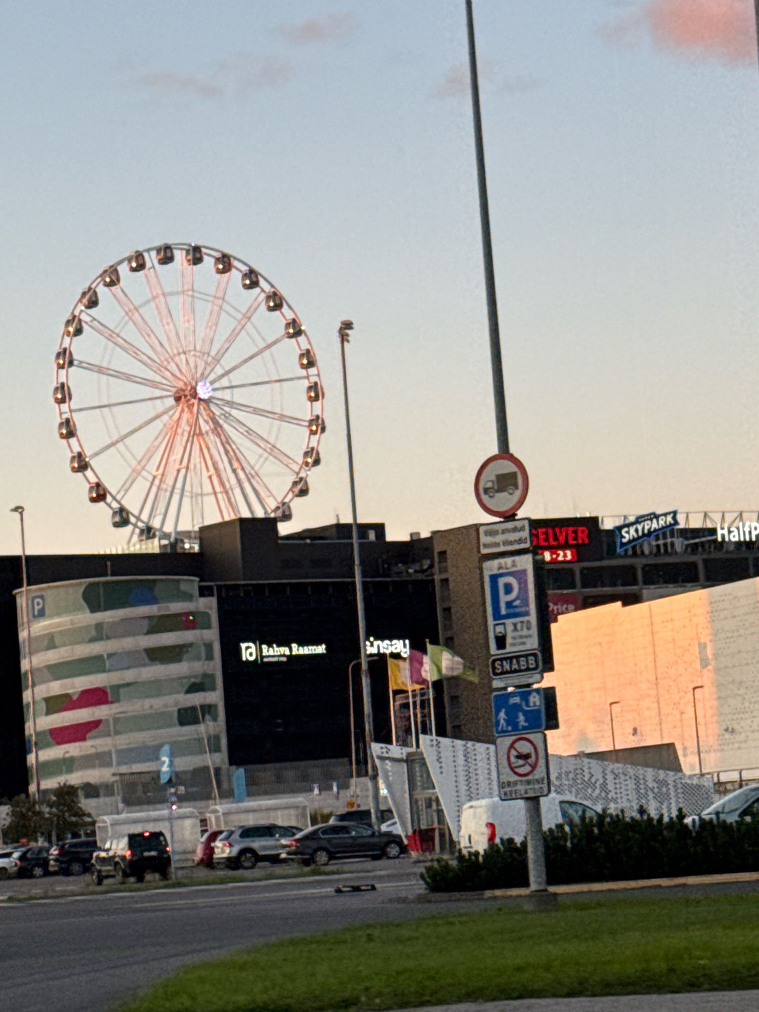 The image shows a Ferris wheel in the background, with a modern building facade and parking lot in the foreground. The Ferris wheel is illuminated with pink and white lights. The building facade features various signs, including "SKYPARK" and "HalfPrice," as well as a sign with a truck symbol indicating a no-trucks zone. There is also a parking sign with the letter "P" and some text in Estonian. In the foreground, several cars are parked in a lot, and a white van is partially visible. The scene appears to be in an urban environment, possibly a shopping center or entertainment complex. The sky is clear with some faint pink clouds. The overall impression is a bright and modern setting. The image was taken in Tallinn, Estonia.