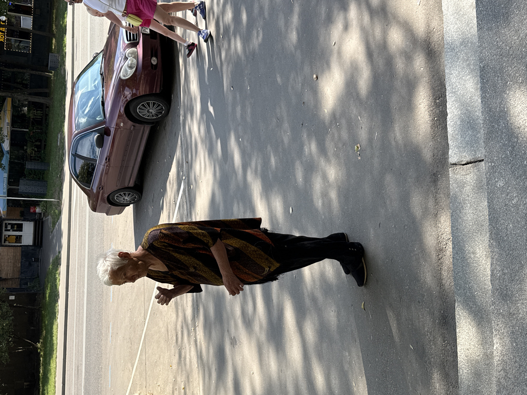 An elderly woman with white hair stands on a sidewalk in Moscow, Russia, wearing a striped poncho and black pants. She is positioned on the right side of the frame, facing left, with her right hand raised and holding a small object. Behind her, a maroon sedan is parked on the street, and two individuals, one in pink shorts, are walking away from the camera in the distance. The scene is illuminated by sunlight, casting dappled shadows on the asphalt. The background features trees and a building with a white door.