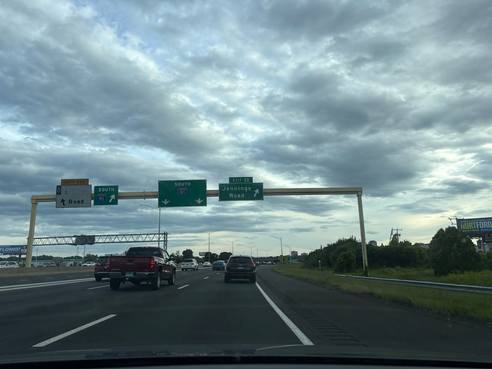 A cloudy day scene shows traffic on a highway in Hartford, Connecticut. Overhead signs indicate "LEFT EXIT" and "SOUTH 91" with an arrow pointing left, and "SOUTH 91" with an arrow pointing up. Another sign indicates "EXIT 33" and "Jennings Road" with an arrow pointing up. Vehicles include a red pickup truck, a black SUV, and several other cars. A billboard on the right side of the road advertises "HURTFORD". The environment is urban with trees and buildings visible in the background.