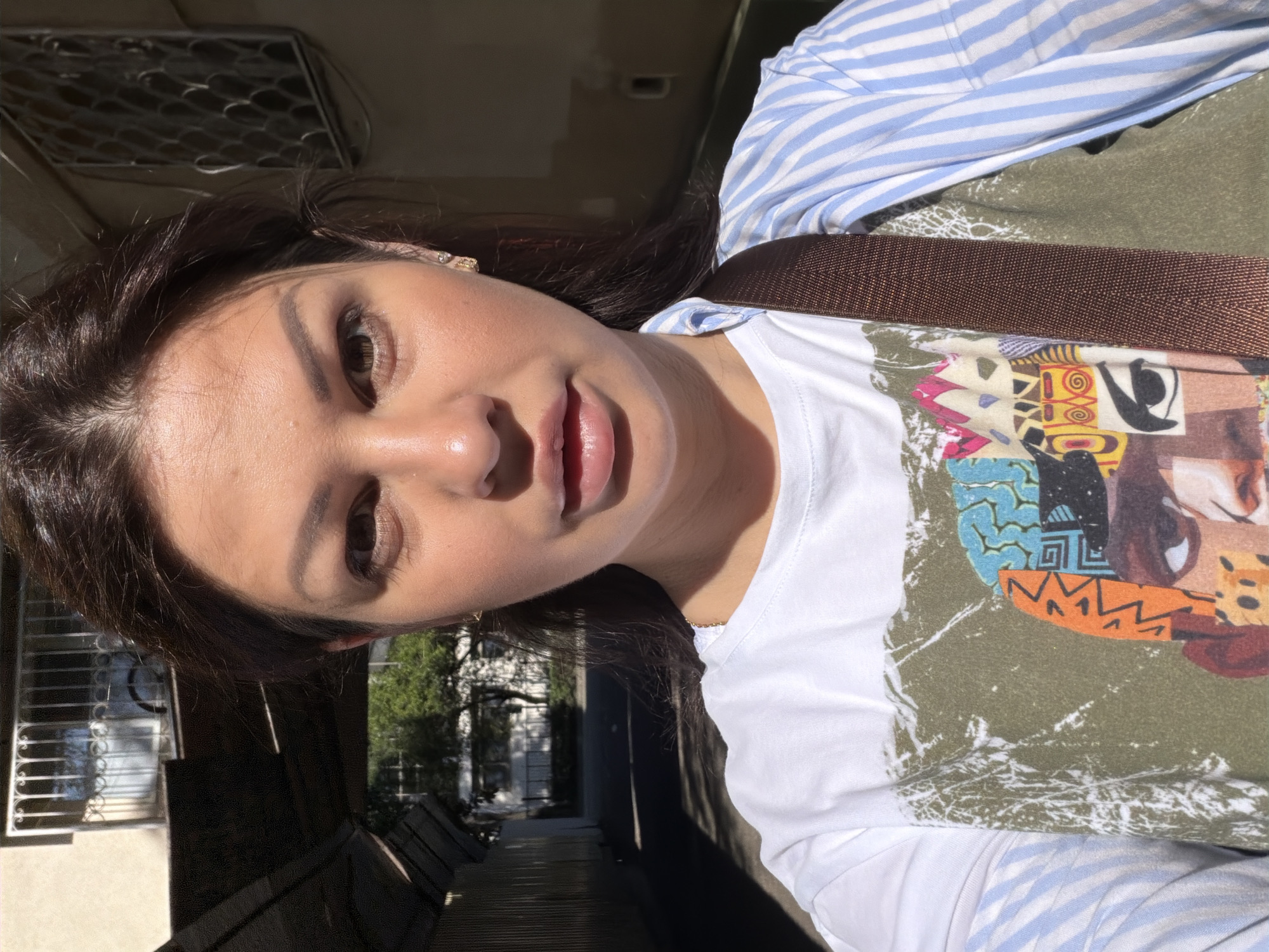 A medium-shot outdoor selfie captures an adult female looking directly at the camera in Sochi, Russia. Her dark hair is pulled back, and her face is illuminated by strong direct sunlight from the viewer's left, casting the right side into shadow. She wears a white t-shirt featuring a multi-colored graphic print and a blue and white striped long-sleeved shirt underneath or layered. A dark brown strap, likely from a bag, diagonally crosses her chest. A small, light-colored stud earring is visible in her right ear. The background depicts an urban environment, likely an alleyway or passage between buildings, with a dark overhead covering structure on the left casting deep shadows. Further back, a sunlit paved area is visible, bordered by trees and light-colored buildings. One building on the upper left features a window with a rectilinear metal grate, and another on the upper right has a window with a patterned metal grate.