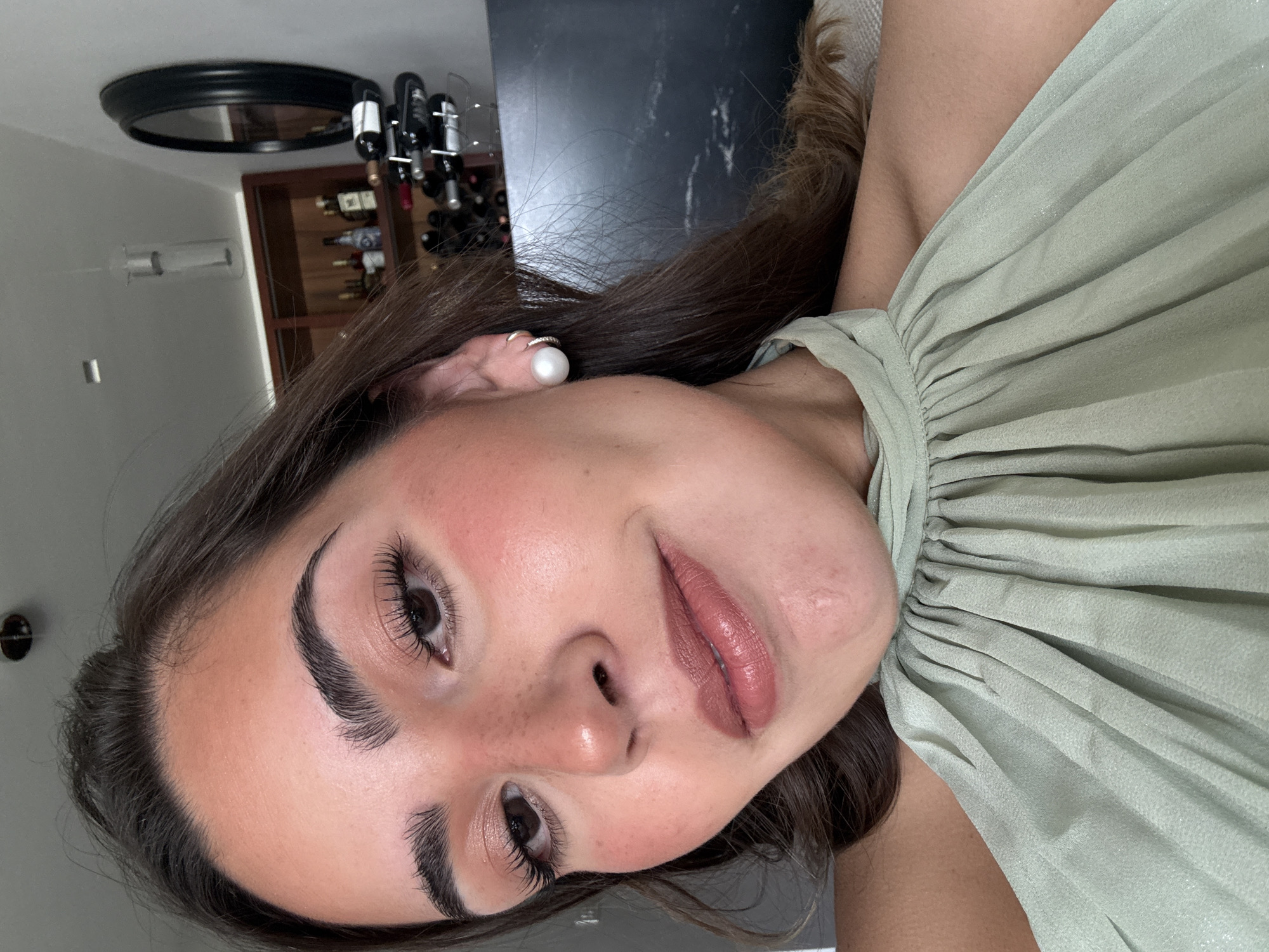 A close-up, rotated selfie depicts a young woman with light skin, brown hair, and dark eyebrows. She is wearing a pearl earring and a light green garment. The background reveals a kitchen area with a black countertop, a wine rack containing several wine bottles, and a round black mirror on the wall. The image does not contain enough information to precisely determine the location within San Bernardino Tlaxcalancingo, Mexico.