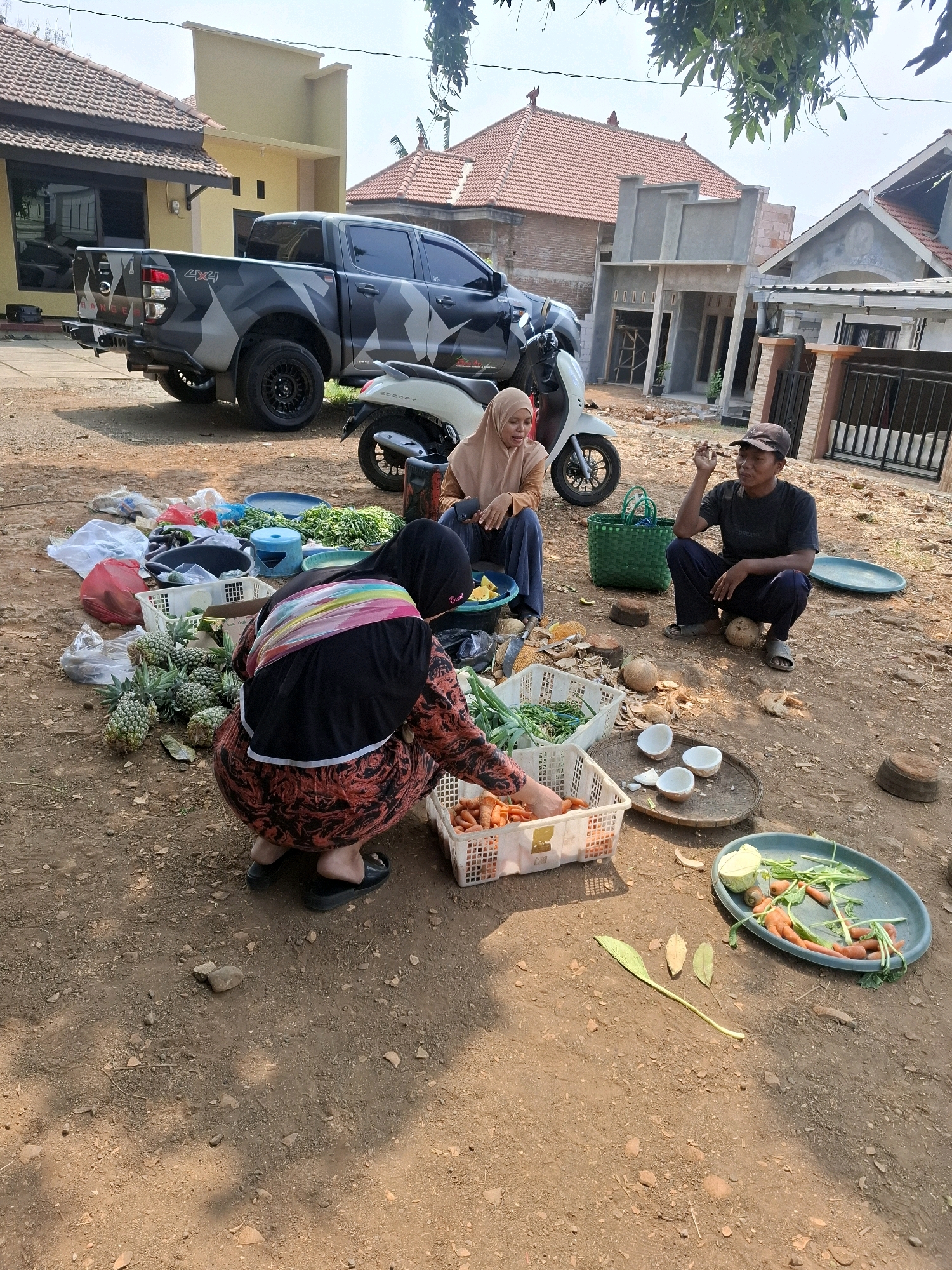 In Dusun Dua, Indonesia, three individuals are present in an outdoor setting with a dirt ground. A dark-colored pickup truck with camouflage detailing and a motorcycle are parked in the background, near some houses. In the foreground, a woman wearing a black hijab and a patterned red and black dress is kneeling and sorting through produce, likely for sale. She is next to a white plastic crate filled with carrots. To her left, another woman in a beige hijab is seated and appears to be looking at her phone. A man in a dark t-shirt and baseball cap is squatting and holding something in his hand, possibly a cigarette. Several crates and baskets containing various vegetables, pineapples, and coconuts are scattered around the area. The environment suggests a rural or semi-rural marketplace or roadside stall.
