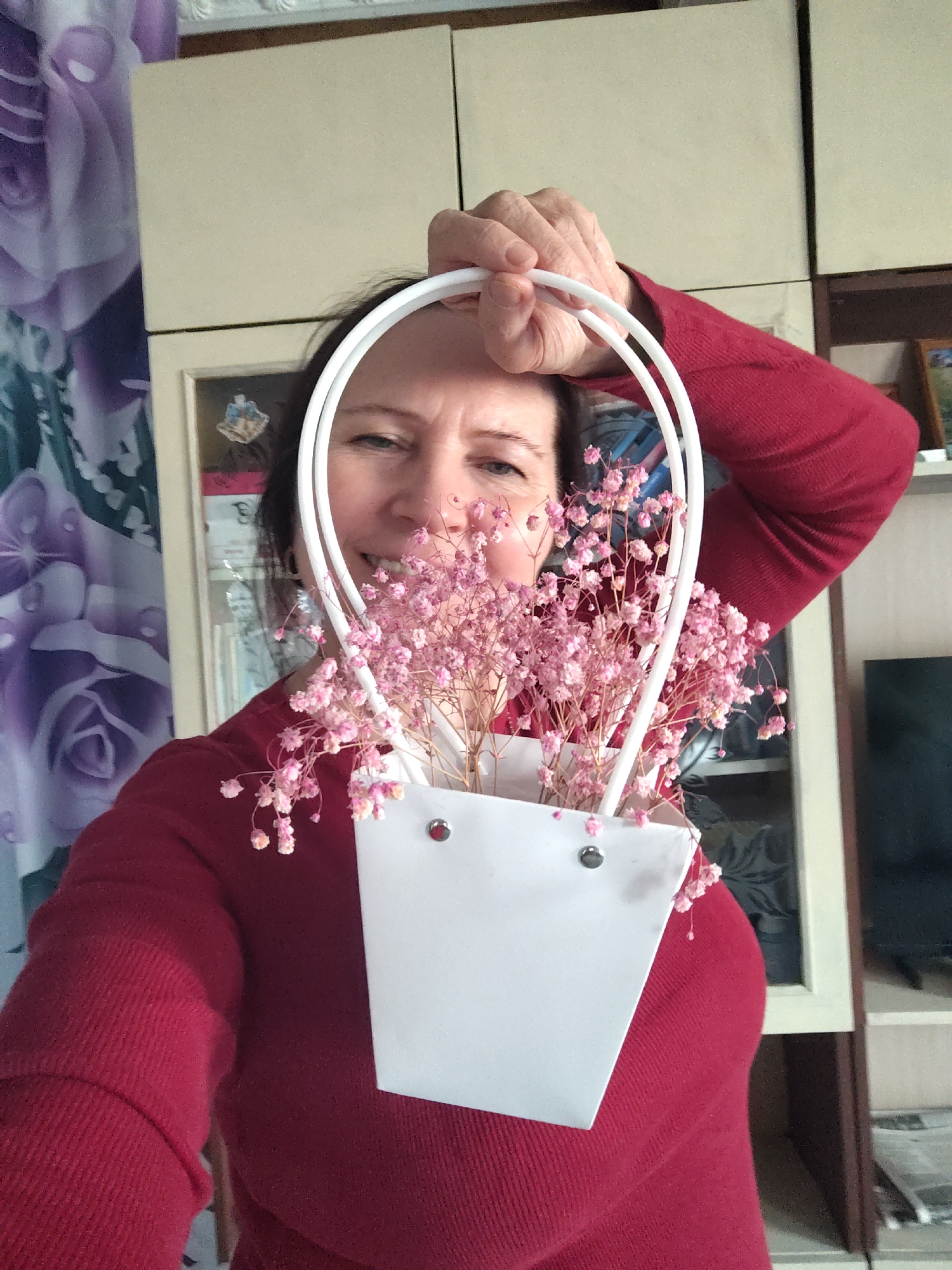 A person, possibly a woman, is holding a white paper bag with pink flowers inside in front of her face. The bag has white handles. The person is wearing a red sweater. The background shows a cabinet and a curtain with a purple rose pattern. A television is also visible in the background. The location is possibly in Сусады-Эбалак, Russia.