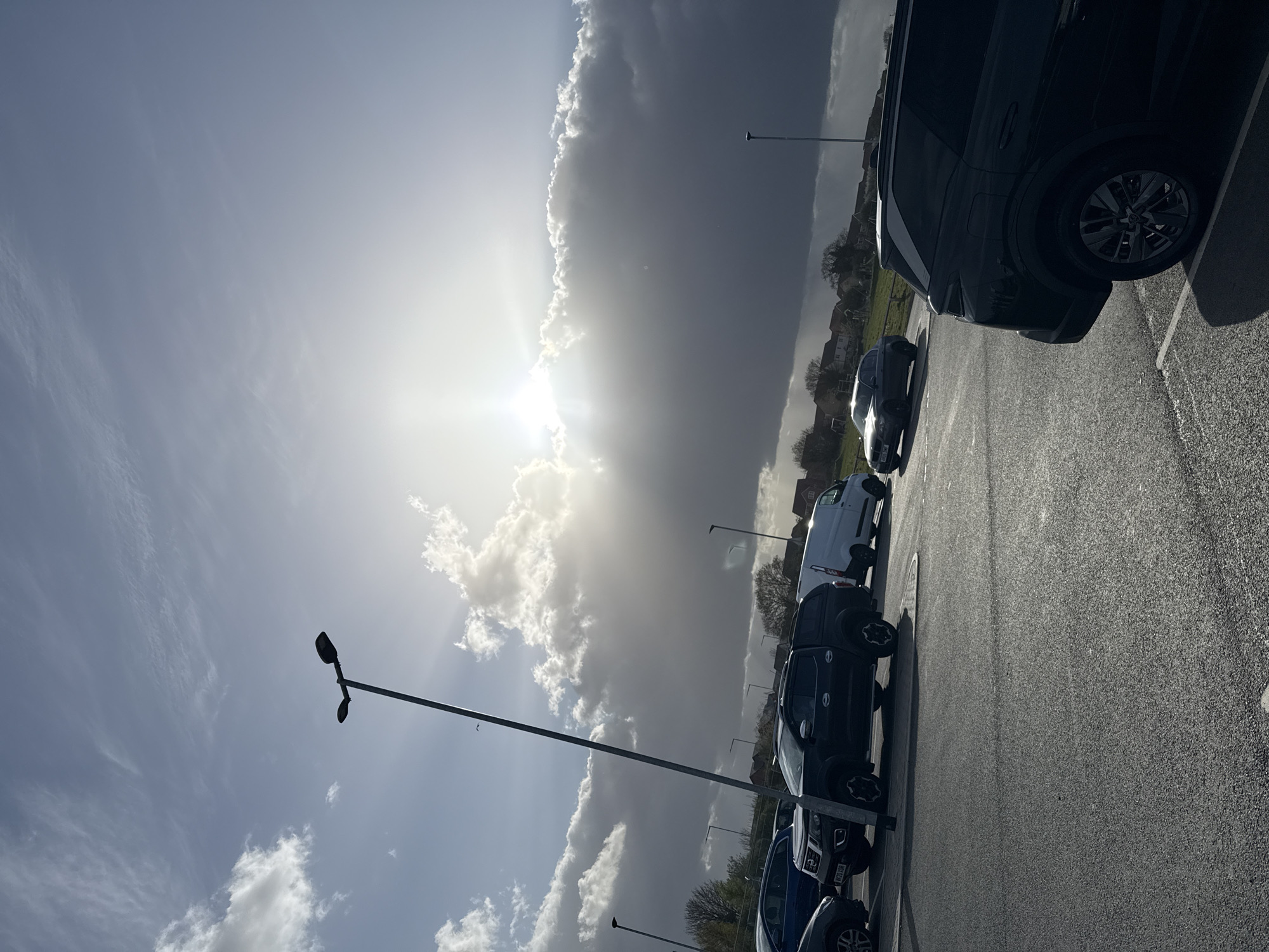 The image shows a parking lot in daylight. Several cars are parked in a row. The sky is partly cloudy, with the sun visible behind the clouds. Street lights are also present. The location is in Great Yarmouth, United Kingdom.