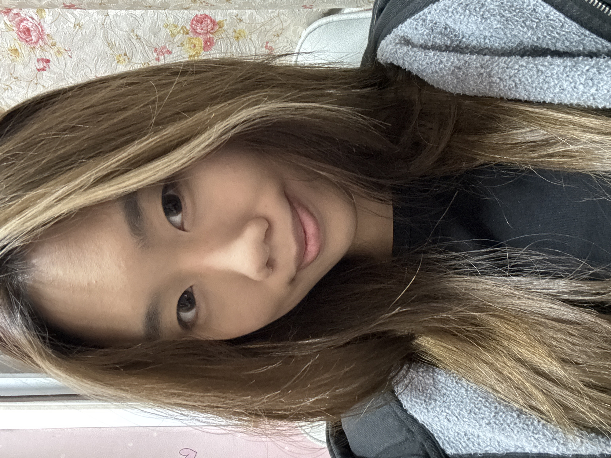 A head-and-shoulders shot of a young woman with long brown hair. She is smiling slightly. She is wearing a black top with a gray hoodie. The background is a light-colored wall with a floral pattern. The specific location in Shapingba District, China, cannot be determined from the image.