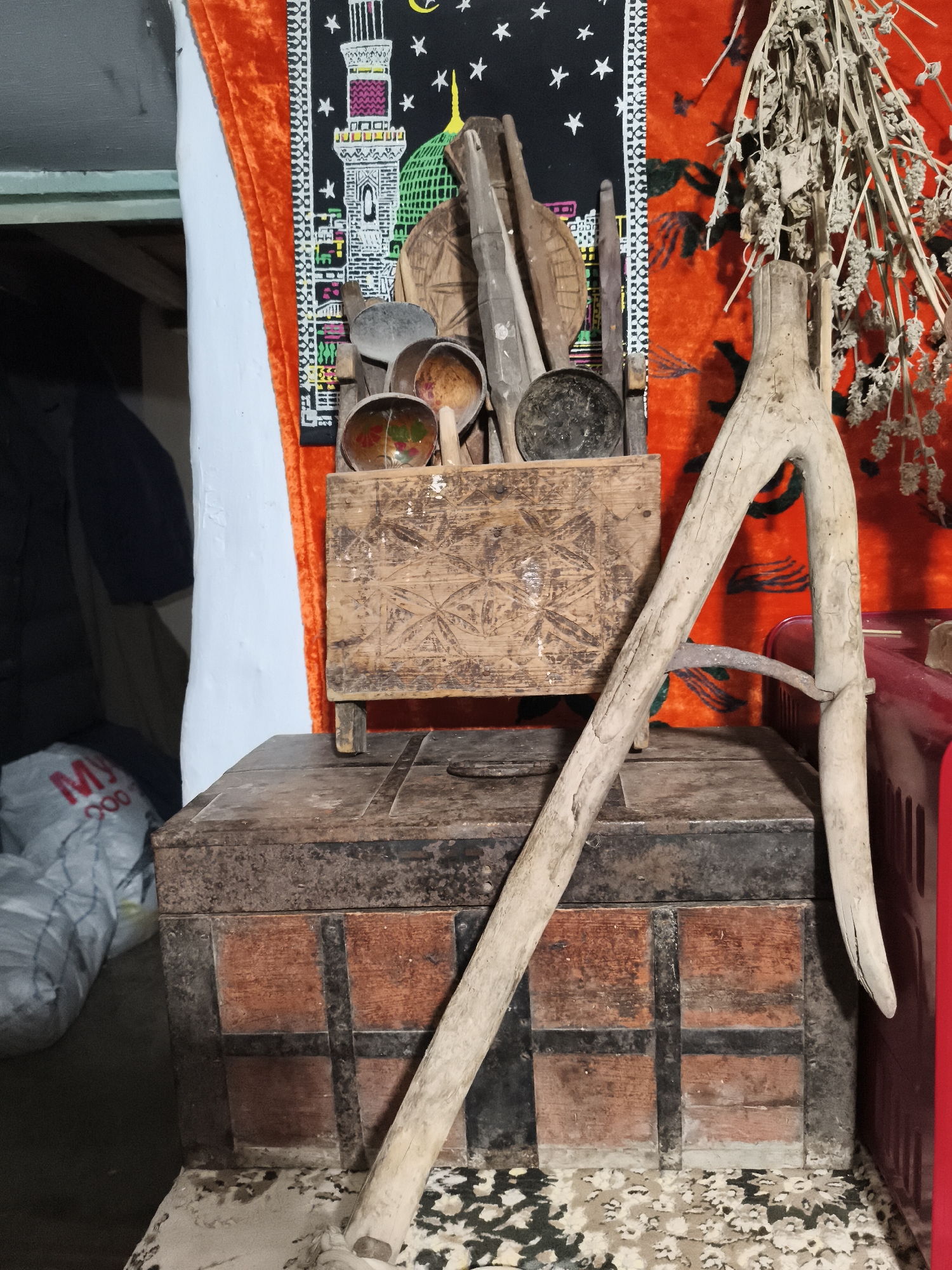 In Irgali, Russia, a dimly lit room contains a collection of rustic objects. Dominating the foreground is a large, dark wooden chest with metal banding. Leaning against it and extending upwards is a light-colored wooden tool, possibly part of an agricultural implement. Resting on top of the chest is a smaller, carved wooden box. Inside the smaller box, several items are visible, including a metal pan and several carved wooden bowls or containers, some of which appear to be made from gourds. Behind these objects, a vibrantly colored tapestry or rug with an intricate pattern hangs on the wall, featuring an image of a mosque and star motifs on a black background. To the right of the tapestry, a red object, possibly a container or piece of furniture, is partially visible. Dried plant material hangs from the top right of the frame. The overall impression is one of a storage area or a display of traditional, possibly antique, household items in a rural setting.