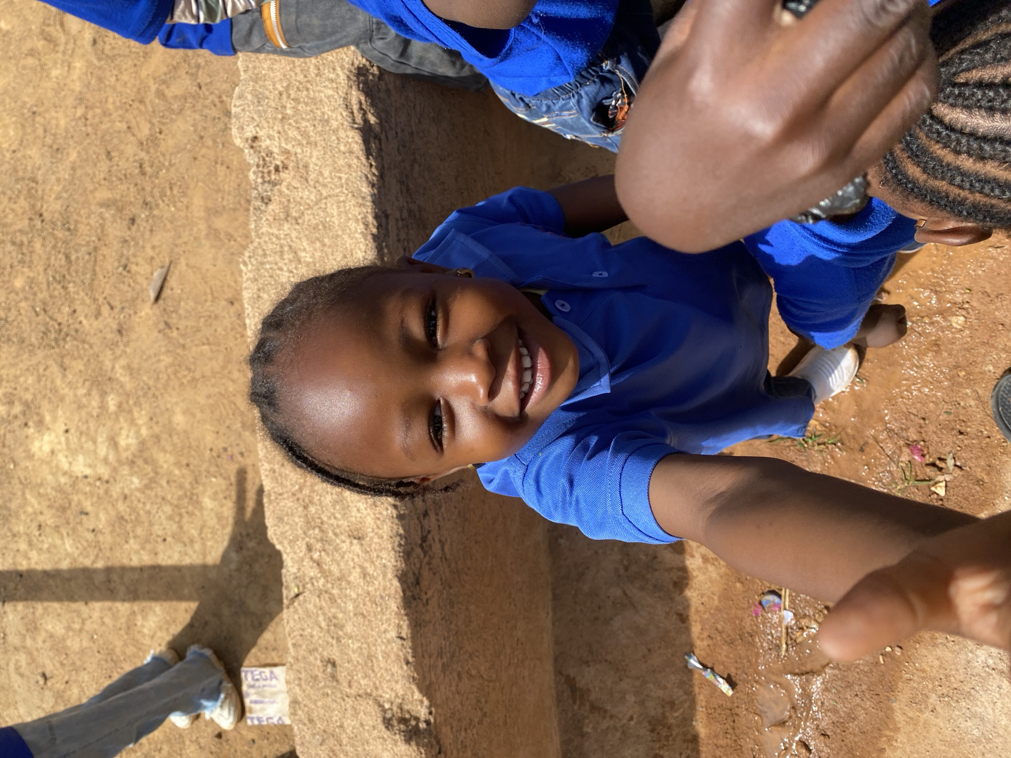 A child with braided hair, wearing a blue collared shirt, smiles directly at the camera, positioned at a low angle. Another child wearing blue and denim is visible in the upper portion of the frame. An adult arm and hand, with a silver ring, is also visible. The ground appears to be a mix of dirt, concrete, and possibly water, with scattered small objects like trash or debris. The scene is outdoors, in what appears to be Gura Top, Nigeria.