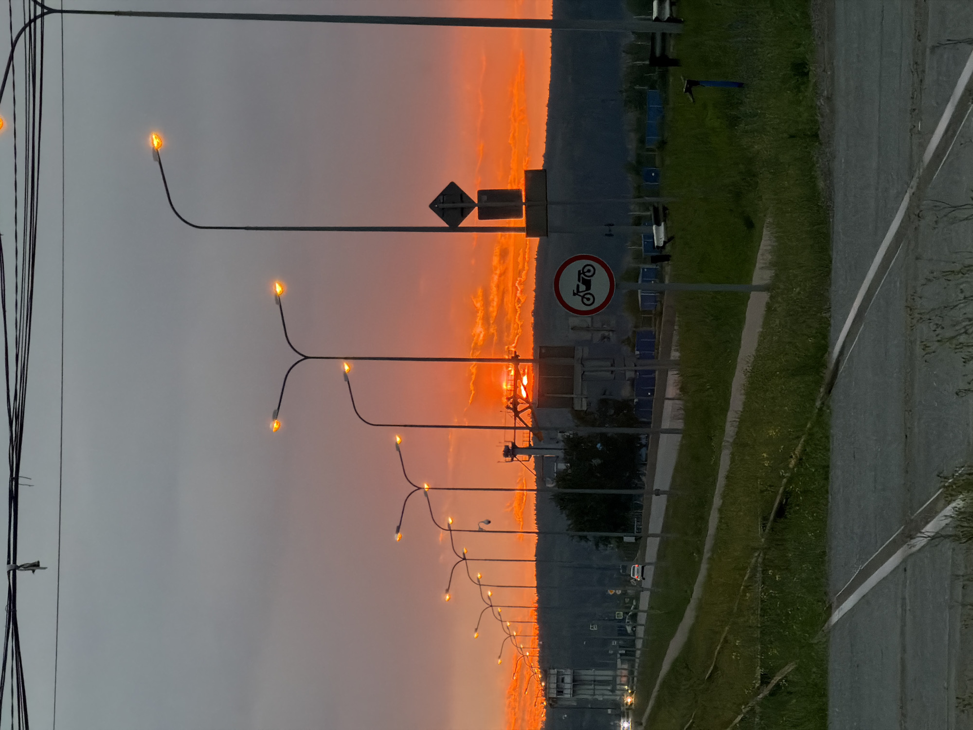 The image depicts a sunset scene on a road in Chaikovsky, Russia. The sky is ablaze with vibrant orange and red hues from the setting sun, with scattered clouds adding texture. A series of streetlights line the road, their lamps glowing, suggesting dusk. In the foreground, train tracks are visible, covered in grass. Further down the road, several cars are driving, and on the right side, there are two road signs, one of which is a circular sign with a motorcycle symbol, indicating a prohibition for motorcycles. Industrial buildings and a tall tower are present in the background, set against a backdrop of a tree-covered hill. The overall atmosphere is transitional, capturing the shift from day to night.