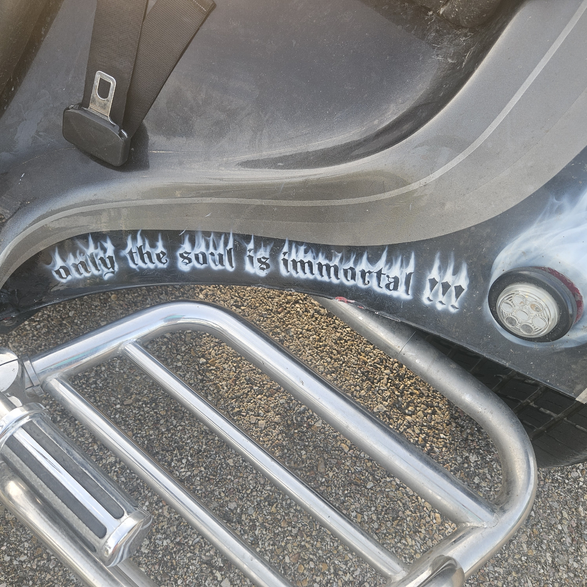The image shows a close-up, eye-level view of the side of a black vehicle, possibly a motorcycle or a custom car, with graffiti that reads "only the soul is immortal" in white, stylized lettering with flame graphics. A silver chrome bumper or footrest is prominent in the foreground, with a dark textured surface of the vehicle behind it. A black seatbelt with a silver buckle is visible at the top left of the frame. The vehicle appears to be parked outdoors on a surface of gravel. The overall condition of the vehicle's surface suggests it may be dusty or worn. No people are present in the image. The location is identified as Illescas, Spain.