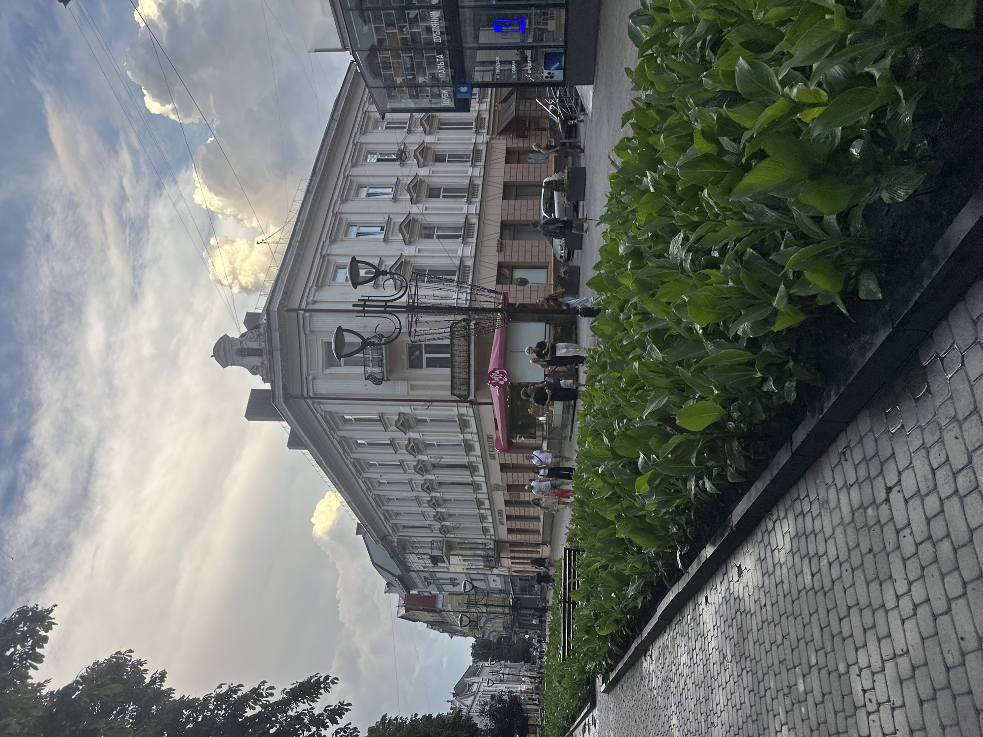 The image depicts a street scene in Ternopil, Ukraine. In the foreground, a paved walkway made of interlocking grey bricks is visible, wet from recent rain. Adjacent to the walkway is a raised flower bed filled with lush, green plants. Beyond the flower bed, a street lined with buildings stretches into the distance. Several people are walking on the sidewalk and crossing the street. The buildings have architectural styles typical of European cities, with ornate facades and multiple stories. One building on the right appears to be a modern retail establishment with large glass windows displaying merchandise. The sky is partly cloudy with dramatic, illuminated clouds, suggesting either sunrise or sunset. A street lamp and a decorative planter are also present in the mid-ground. The overall impression is of a lively urban environment on a somewhat overcast day.