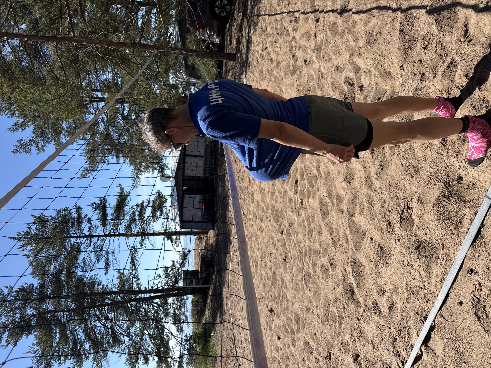 The image captures an outdoor scene at a sandy beach volleyball court in Zelenaya Roshcha, Russia, under a clear blue sky. A male individual, viewed from a slightly elevated rear-left perspective, stands on the court. He is wearing sunglasses, a blue short-sleeved polo shirt with the Cyrillic text "ЦНИ РТ" printed in white on the back, olive green shorts, and pink and black patterned sand socks or shoes. His left arm is bent, and his shirt appears to be pulled or bunched up at the front, suggesting an action of adjustment or holding an object. The black mesh volleyball net with a white top band spans across the middle ground. In the foreground, a white boundary tape for the court is visible on the light brown sand, which is textured with footprints. The background features a body of water, consistent with the Gulf of Finland or a large lake, bordered by numerous tall pine trees. Several small, utilitarian structures and a dark-colored SUV are partially visible behind the trees to the right. The scene is brightly lit by direct sunlight, casting distinct shadows on the sand.
