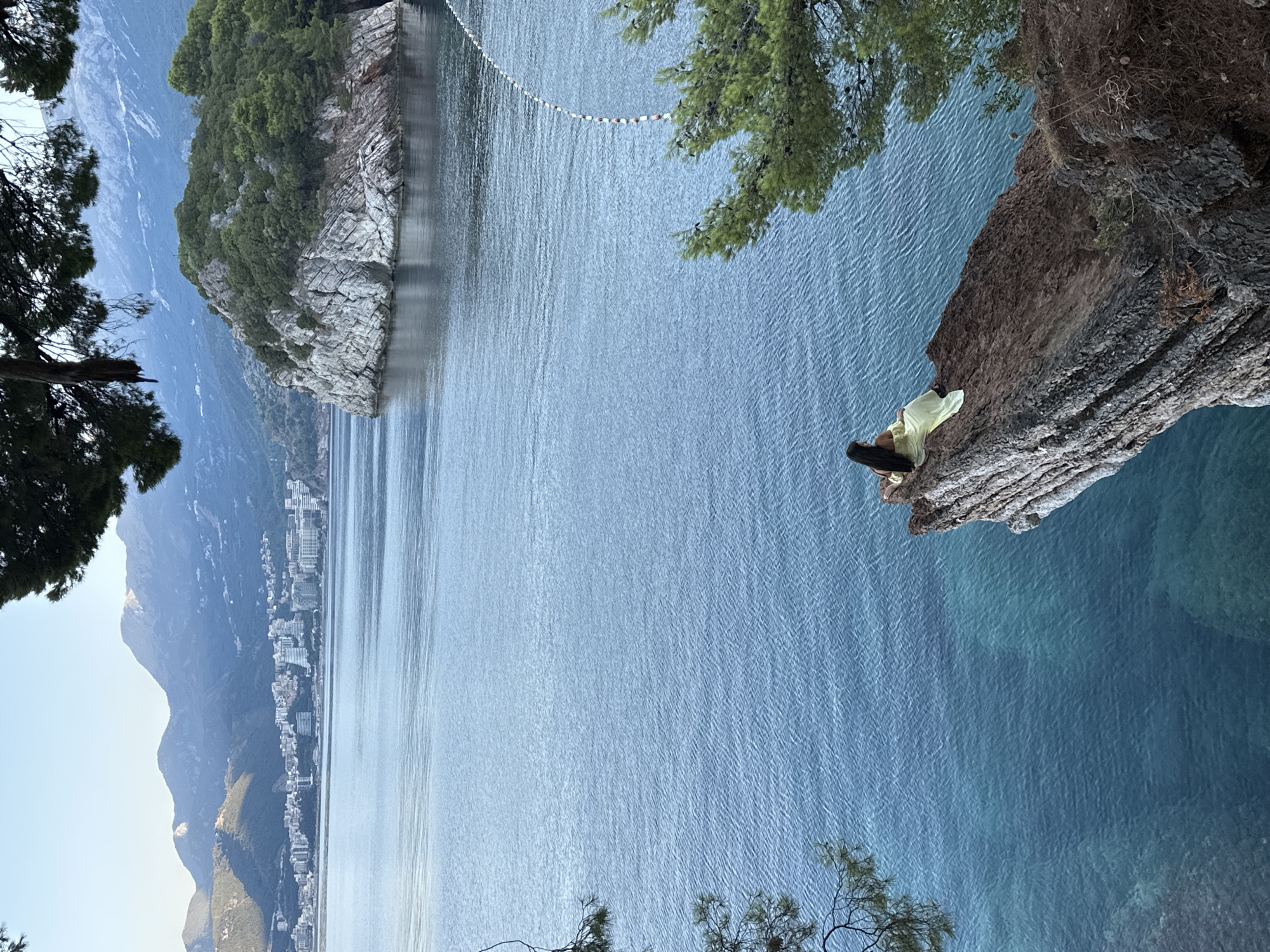 A person wearing a yellow dress is sitting on the edge of a cliff overlooking a body of water. The person is facing away from the camera, looking out at the scenery. In the background, there is a town with buildings and mountains. The water is a deep blue, with some lighter shades indicating shallower areas. There are trees on the cliff and in the background. The sky is clear and pale blue. The image appears to be taken from a high vantage point. The location is the coast near Sveti Stefan, Montenegro, a picturesque resort town known for its island hotel.