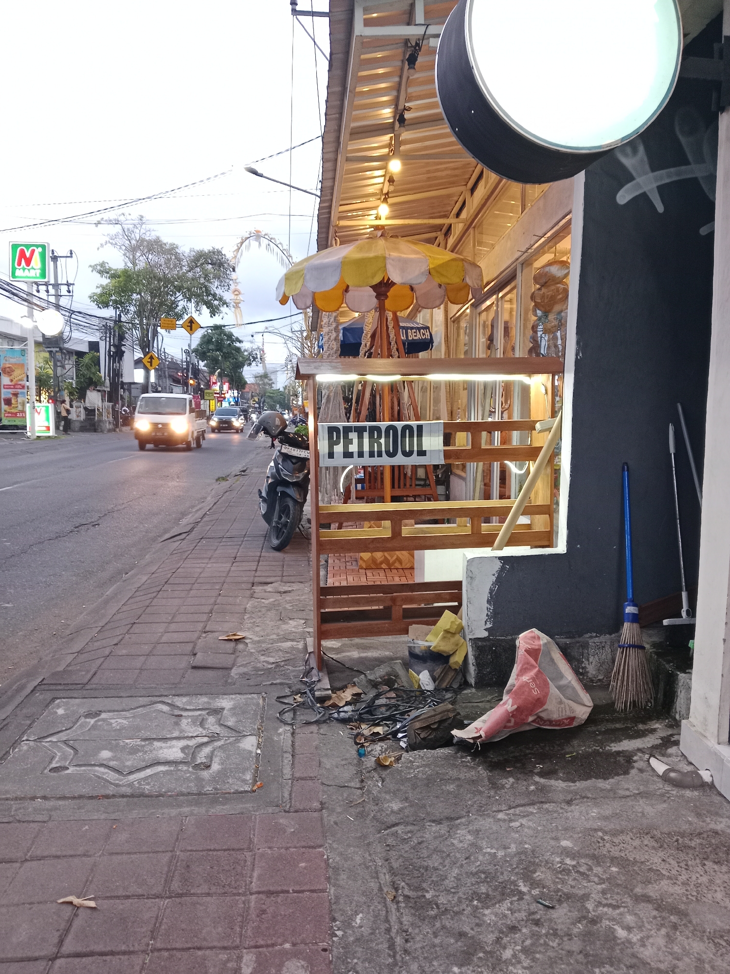 The image depicts a street scene in North Kuta, Indonesia, likely at dusk. The foreground shows a paved sidewalk with a patterned manhole cover and some debris, including a plastic bag with red and white markings, a broom with a blue handle, and another broom leaning against a building. Behind the sidewalk, a low wooden structure displays a sign that reads "PETROOL". A motorcycle is parked to the left of this structure. The street itself is paved and appears to be a busy road, with a white utility vehicle and a car visible in motion. Several traffic signs and streetlights line the road. Shops with illuminated awnings and displays are visible on the right side of the street. A large, round, illuminated sign hangs from the awning of one of the shops. Trees and power lines are present in the background. The sky is overcast, suggesting a cloudy or twilight atmosphere.