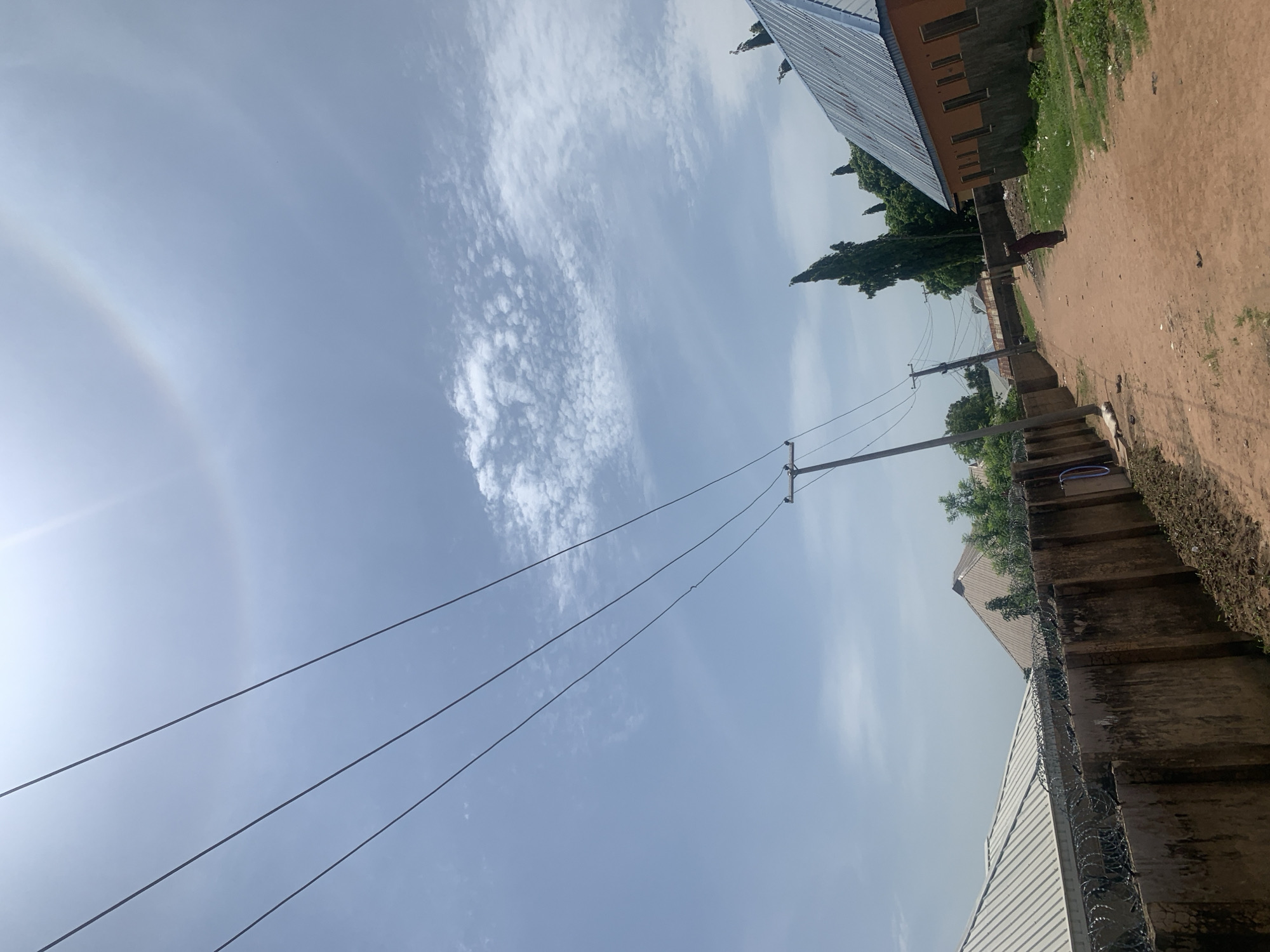 The image depicts a dusty, unpaved street in Jauro Yinu, Nigeria, under a bright, partly cloudy sky. Power lines stretch across the frame, supported by utility poles. On the left, a tall, concrete wall topped with razor wire runs along the street, with corrugated metal roofs of buildings visible behind it. On the right, an orange building with a blue metal roof stands next to a patch of green grass and scattered debris. A single person, dressed in dark clothing, walks away from the camera down the street. In the sky, a faint rainbow or solar halo is visible.