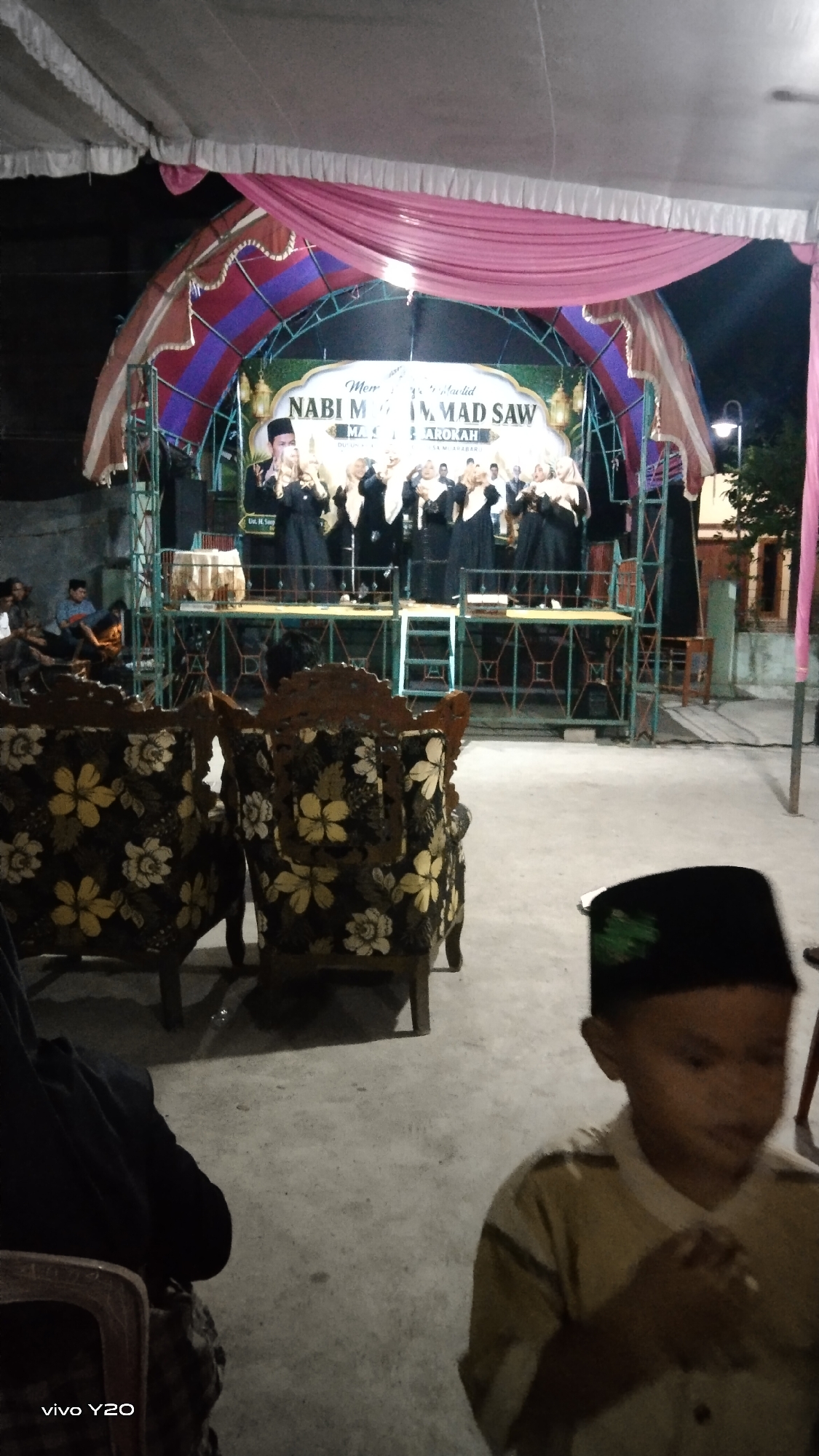 An outdoor stage event is captured at night in Karawang, Indonesia, likely a religious gathering or celebration, indicated by the banner text "NABI MUHAMMAD SAW". The stage, adorned with pink and purple draping, features a group of individuals, likely performers, dressed in black attire and headscarves, standing on a raised platform. Several chairs with floral upholstery are positioned in the foreground, suggesting an audience seating area. In the lower right corner, a young boy wearing a black cap and a light-colored garment is in focus, seemingly observing the performance. The ground appears to be a concrete surface, and the overall lighting suggests artificial illumination is in use for the event.