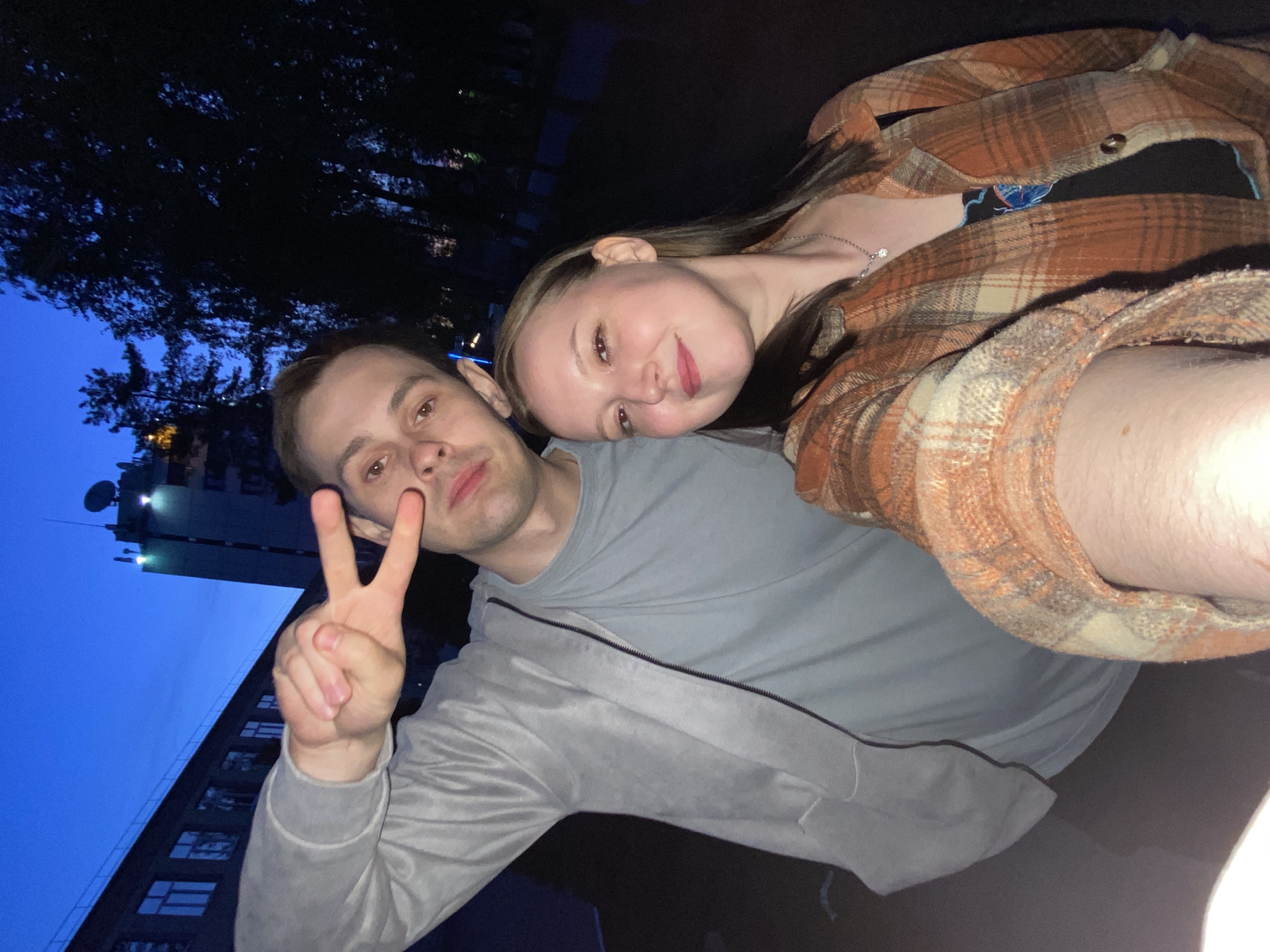 A selfie was taken at night by a male and a female in front of a building. The location appears to be in Chaikovsky, Russia, given the architectural style of the building, which features visible communication equipment on the roof. The male, wearing a grey jacket, is making a peace sign with his right hand. The female, wearing an orange plaid shirt, is leaning her head onto the male's shoulder. The background features a dark sky with trees and the mentioned building.