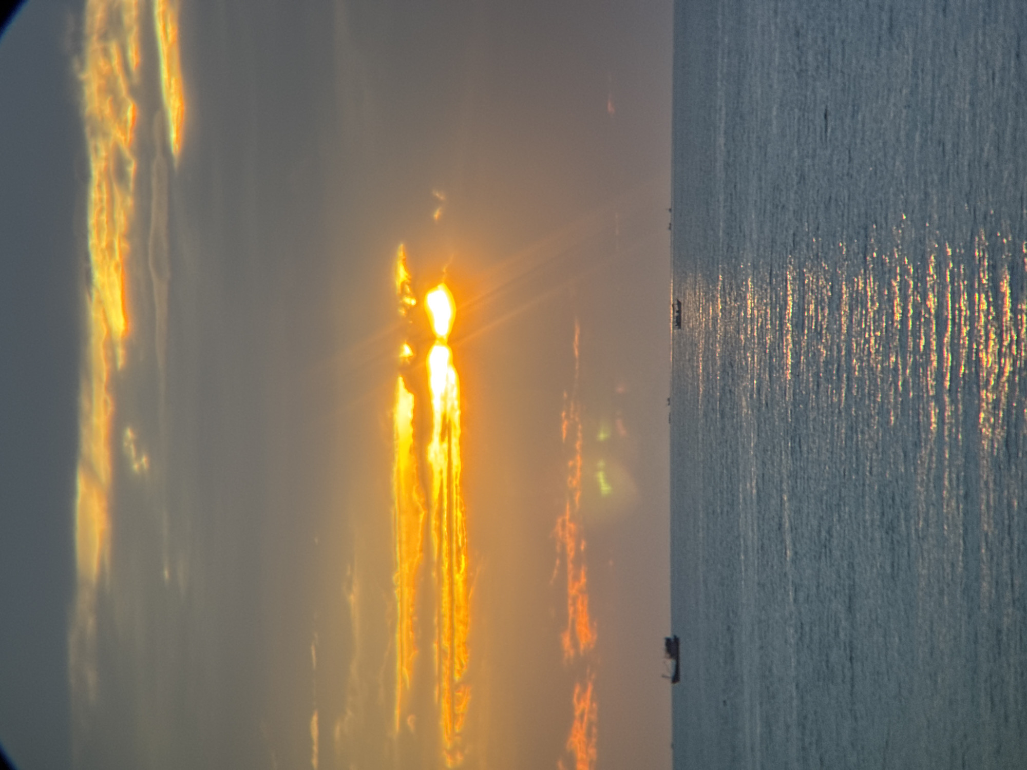 The image depicts a sunset over the ocean in Nha Trang, Vietnam. The sun, partially obscured by clouds, casts a warm orange glow across the sky and reflects on the rippling surface of the water. Several boats are visible on the horizon, appearing as small silhouettes against the vast expanse of the sea. The sky is a gradient of dark gray at the top, transitioning to lighter shades of orange and yellow closer to the horizon, with streaks of sunlight breaking through the clouds. The water is a deep blue, with shimmering reflections of the sunset scattered across its surface.