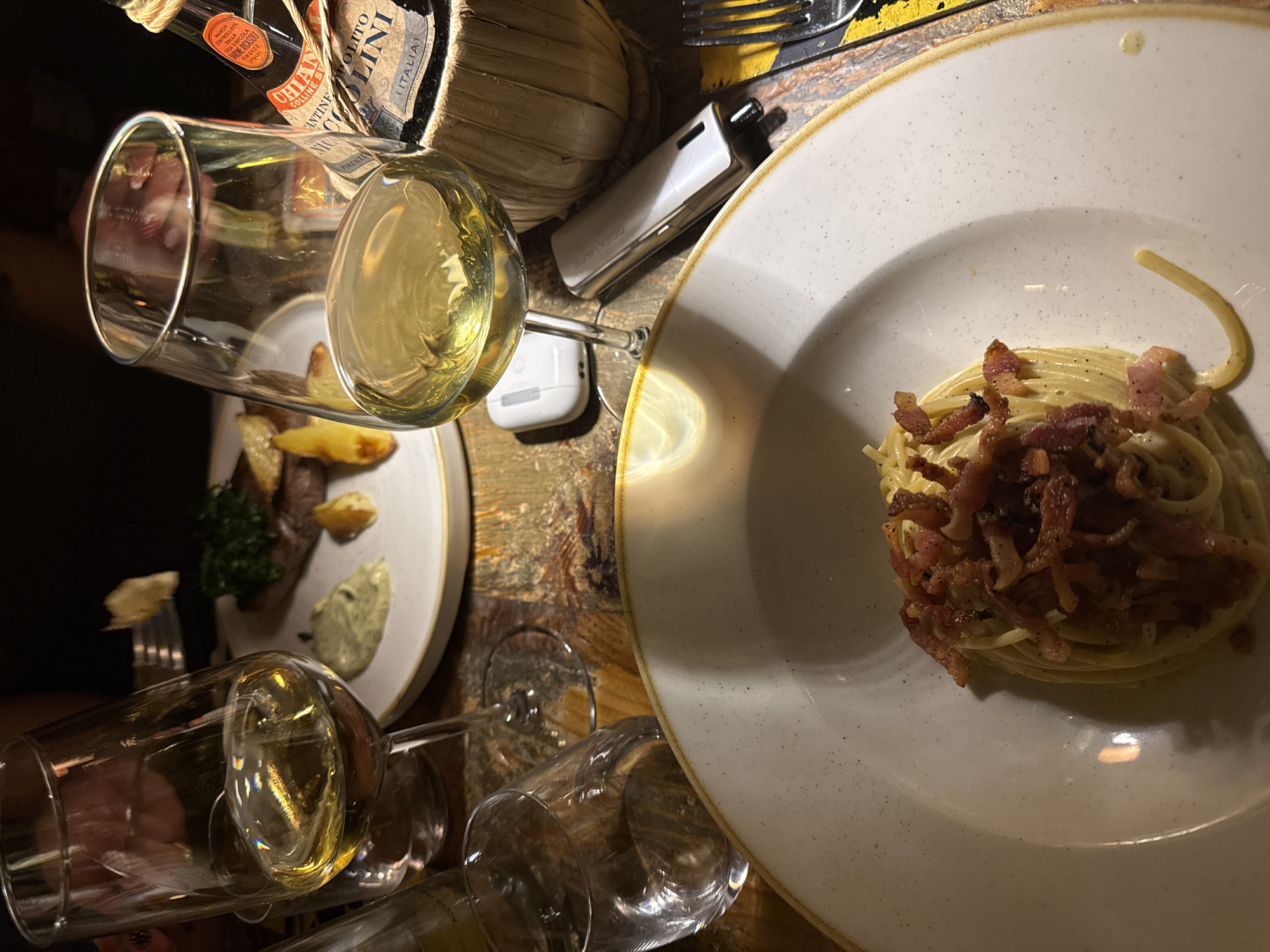 The image depicts an indoor dining scene at an Italian restaurant, consistent with establishments found in Milan, Italy. The environment is dimly lit, showcasing a distressed wooden table surface. In the foreground, a white, speckled ceramic plate presents a serving of spaghetti carbonara, generously topped with crispy bacon or pancetta. To the right, a stemmed wine glass containing a pale yellow liquid, identified as white wine, rests on the table. A white Apple AirPods case is positioned beneath this glass, next to a silver-toned Geekvape vaping device. A silver fork is partially visible on the far right, resting on a dark patterned placemat.
In the mid-ground, a second stemmed wine glass also holds white wine, situated to the left. An empty, smaller water glass is positioned near its base. In the background, a second white, speckled ceramic plate displays a steak, accompanied by roasted potato wedges, a small pile of green garnish, and two distinct sauces. A human hand, adorned with dark nail polish, holds a fork with a piece of food over this steak, indicating active consumption. A Chianti wine bottle, distinctively wrapped in a straw fiaschetto and labeled "ANTINE S. POLITO CELLINI (ITALIA)," stands on the right side of the table, behind the right wine glass. The scene implies a social dining interaction among at least two individuals.