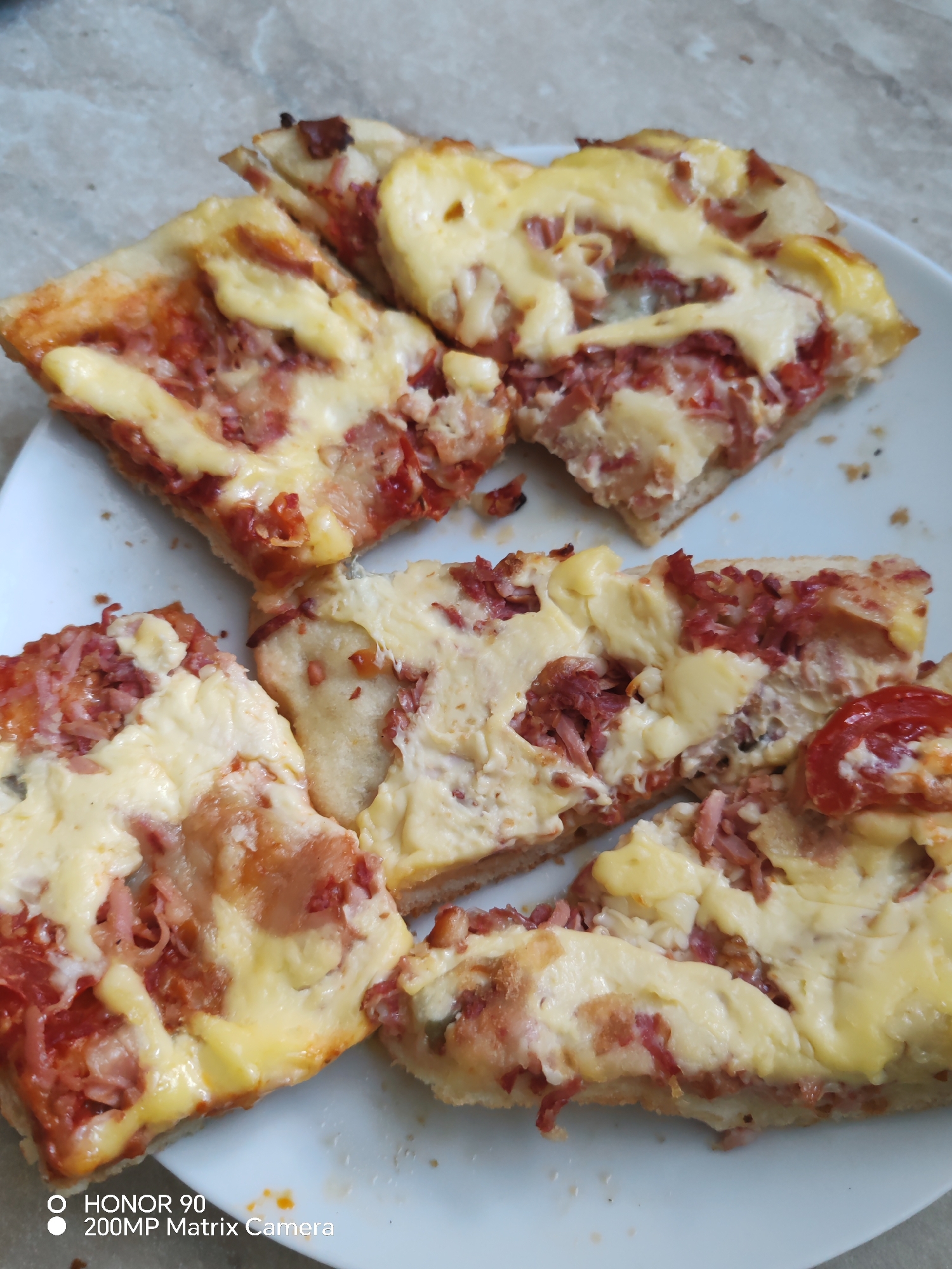 The image shows several slices of pizza arranged on a white plate. The pizza has a crust, tomato sauce, a generous amount of melted cheese, and visible pieces of meat, likely ham or bacon, and possibly some tomato slices. The pizza appears to be homemade or from a casual eatery, with uneven crust edges and toppings spread across the surface. The background is a textured surface, possibly a countertop or table, with a light brown and gray marble pattern. There is no visible indication of the city of Krasnaya Polyana, Russia, or any people in the image. The bottom left corner of the image contains a watermark that reads "HONOR 90 200MP Matrix Camera".