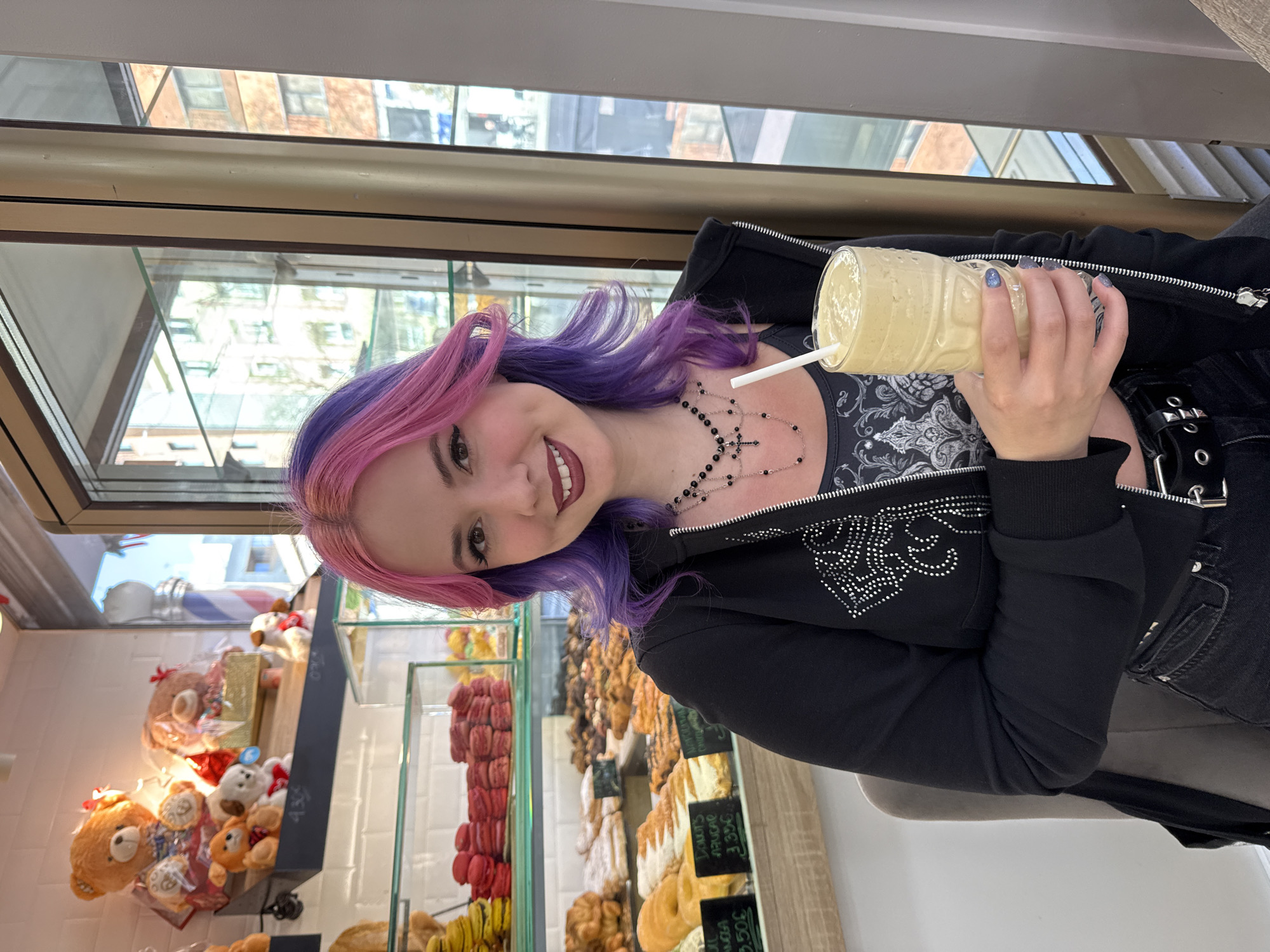 A woman with purple and pink hair is holding a full beverage container with a straw. She is wearing a black jacket, a necklace, and a top with white embellishments. Behind her, a display case shows pastries such as donuts, macarons, and breads, and a shelf presents teddy bears. The window reveals buildings in the background. Based on the presence of goods and architecture visible behind the woman, the location appears to be a bakery or cafe in Madrid, Spain.