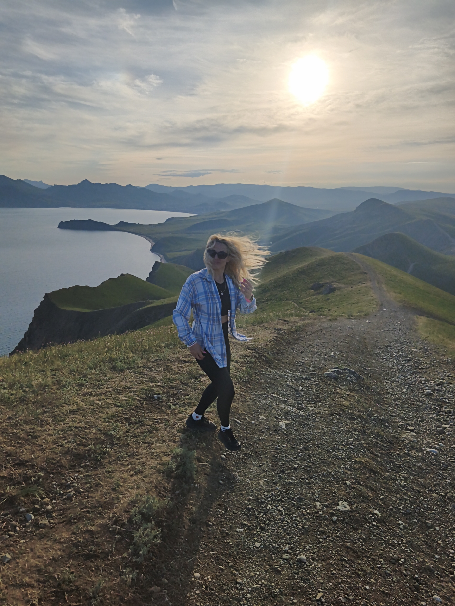 A blonde woman in sunglasses, a blue plaid shirt, black leggings, and black sneakers walks along a ridge on a sunny day. Her hair is blowing in the wind. The ridge is grassy on one side and rocky on the other, with a dirt path running along the top. In the background, there is a body of water with a curved shoreline, and beyond that, rolling hills and mountains fade into the distance. The sun is high in the sky, casting a soft light on the scene. The location appears to be a mountainous region, potentially near the city of Ordzhonikidze, Ukraine.