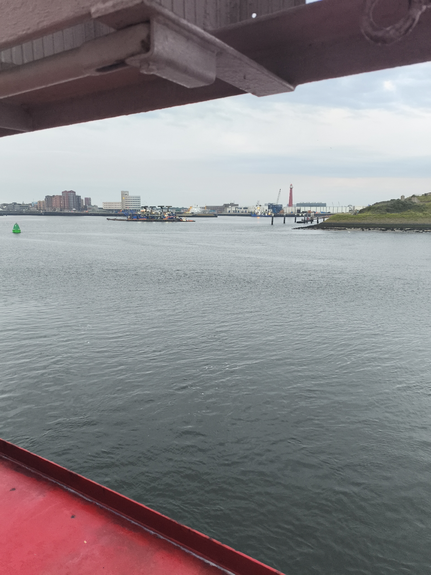 The image captures a waterfront scene in Velsen-Noord, Netherlands, looking across a body of water towards a distant cityscape and industrial area. In the foreground, the weathered, reddish-brown metal structure of a bridge or overhead walkway partially obstructs the view. Below this structure, the calm, grey water of the IJmuiden harbor reflects the overcast sky.
Further across the water, a green navigational buoy is visible to the left. In the middle distance, a collection of barges and vessels are moored, suggesting port activity. Behind these, the skyline of Velsen-Noord is marked by a row of multi-story buildings, some appearing residential and others more industrial. A prominent red lighthouse stands out against the horizon, along with cranes and other port infrastructure. To the right, a grassy, rocky outcrop marks the edge of the land. The overall atmosphere is one of industrial maritime activity under a cloudy sky.
