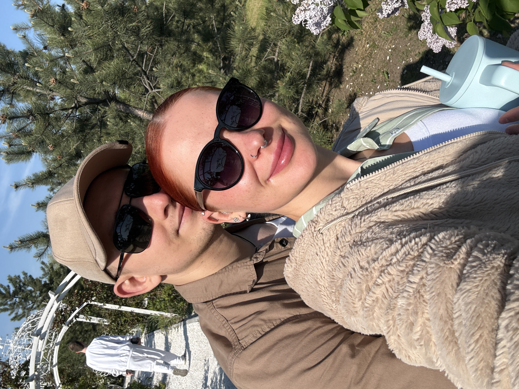 Two people are closely positioned together in an outdoor setting, likely a park or garden. One individual is wearing a cap and sunglasses, while the other is wearing sunglasses and a fuzzy jacket. A light blue cup with a straw is visible. There's a white trellis structure and another person dressed in white in the background. The location is Rostov-on-Don, Russia.