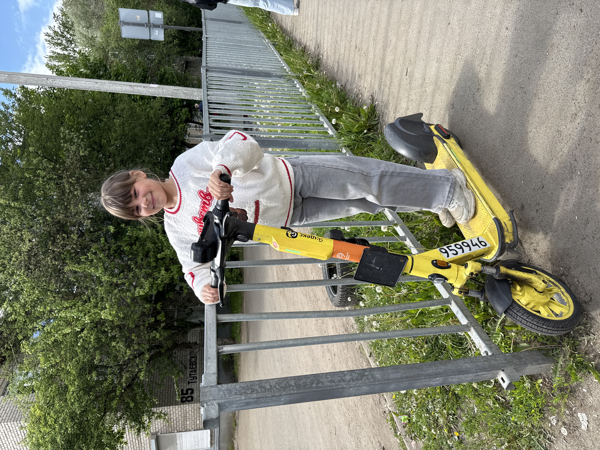 A young girl is standing next to a yellow electric scooter. The scooter appears to be leaning against a metal fence. The fence is located on a sidewalk next to a grassy area with dandelions. A building is behind the fence and trees. There is a sign with "85 ТУТАЕВСК" on the fence. "959946" is written on the scooter. The location is in Шебунино, Russia, based on the sign on the fence.