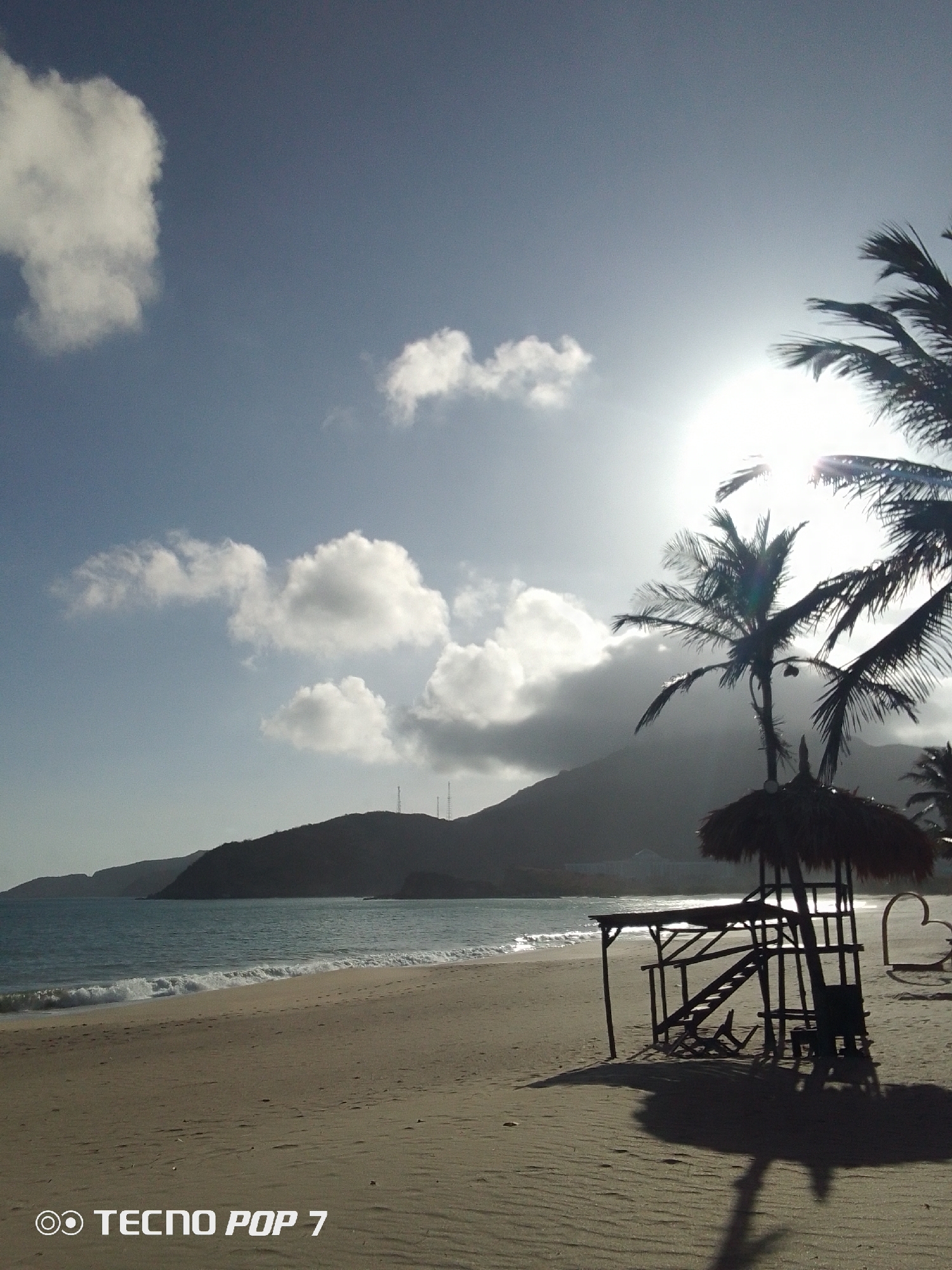 The image depicts a beach scene in Pedro González, Venezuela, under a bright, sunlit sky with scattered clouds. The foreground features a sandy beach with gentle waves lapping the shore. A lifeguard tower, constructed of wood and topped with a thatched roof, stands prominently on the beach, casting a long shadow. Several palm trees are visible near the lifeguard tower. In the distance, a mountain range silhouettes against the sky. The "TECNO POP 7" watermark is at the bottom left corner.