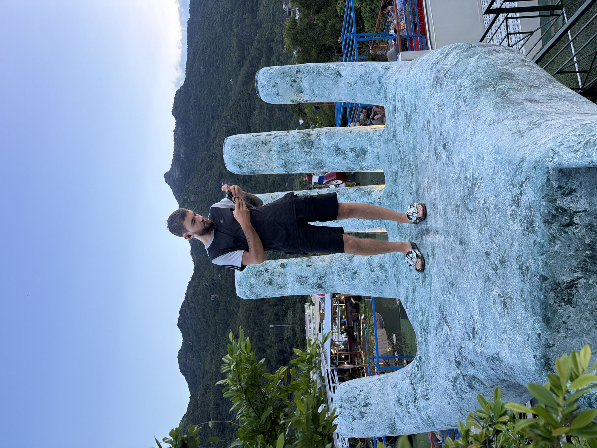 A man stands on a giant hand sculpture, looking at his phone. He is wearing a black t-shirt with gray trim and black shorts, and black and white sandals. The hand sculpture is light blue and textured, with four large finger-like structures rising from the palm. In the background, there are green mountains, a clear blue sky, and some buildings with lights. A tree with green leaves is in the foreground on the left. The location is the Giant Hand, located in Nam Nha Trang Ward, Vietnam.