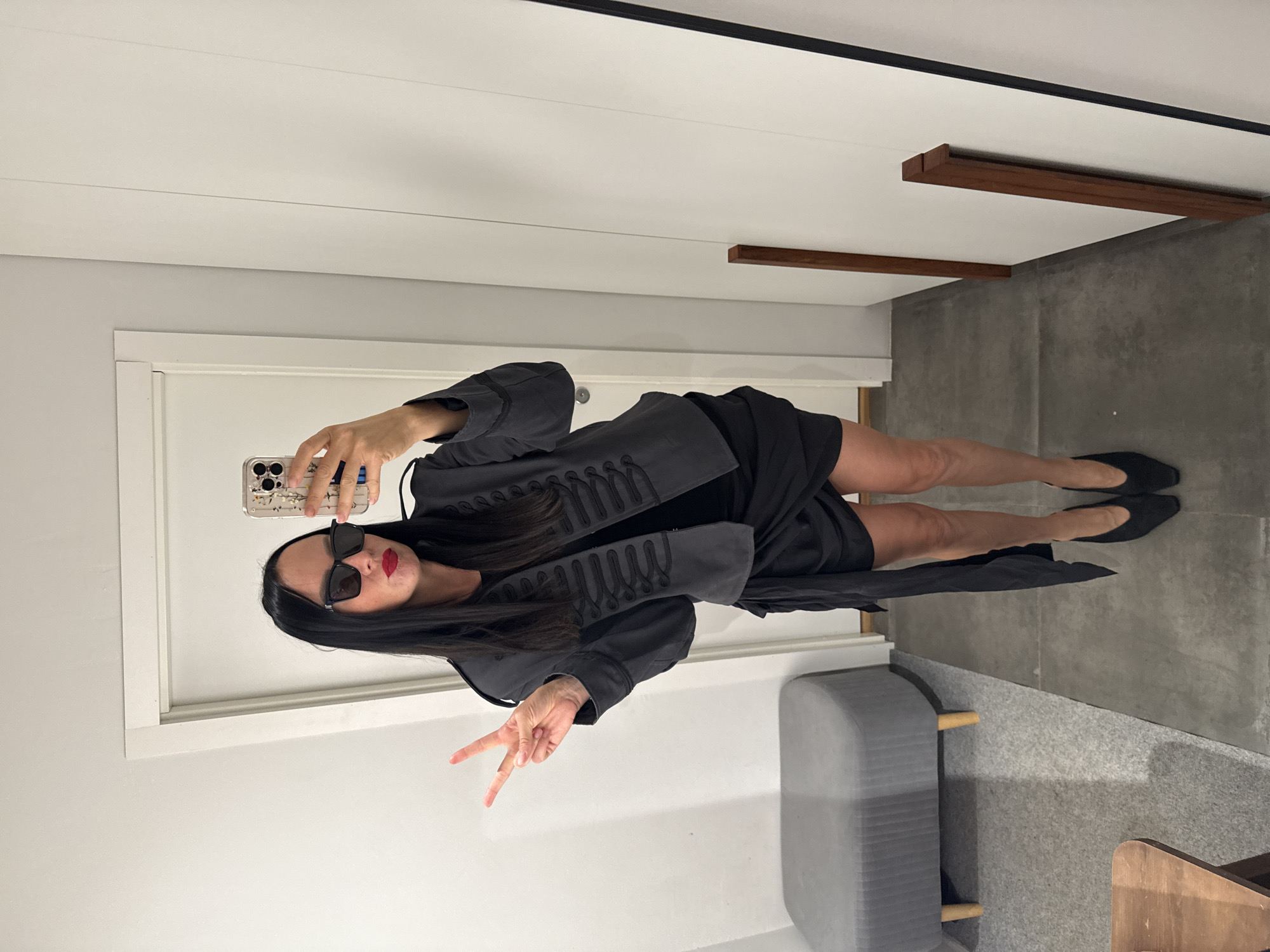 A woman with long dark hair and sunglasses is taking a mirror selfie in a room with white walls and a gray floor. She is wearing a dark gray military-style jacket over a black dress and black high heels. She is holding up two fingers in a peace sign. To her left is a gray upholstered bench and to her right is a tall mirror. There is a wooden table in the foreground. The location is not identifiable as Chaikovsky, Russia.
