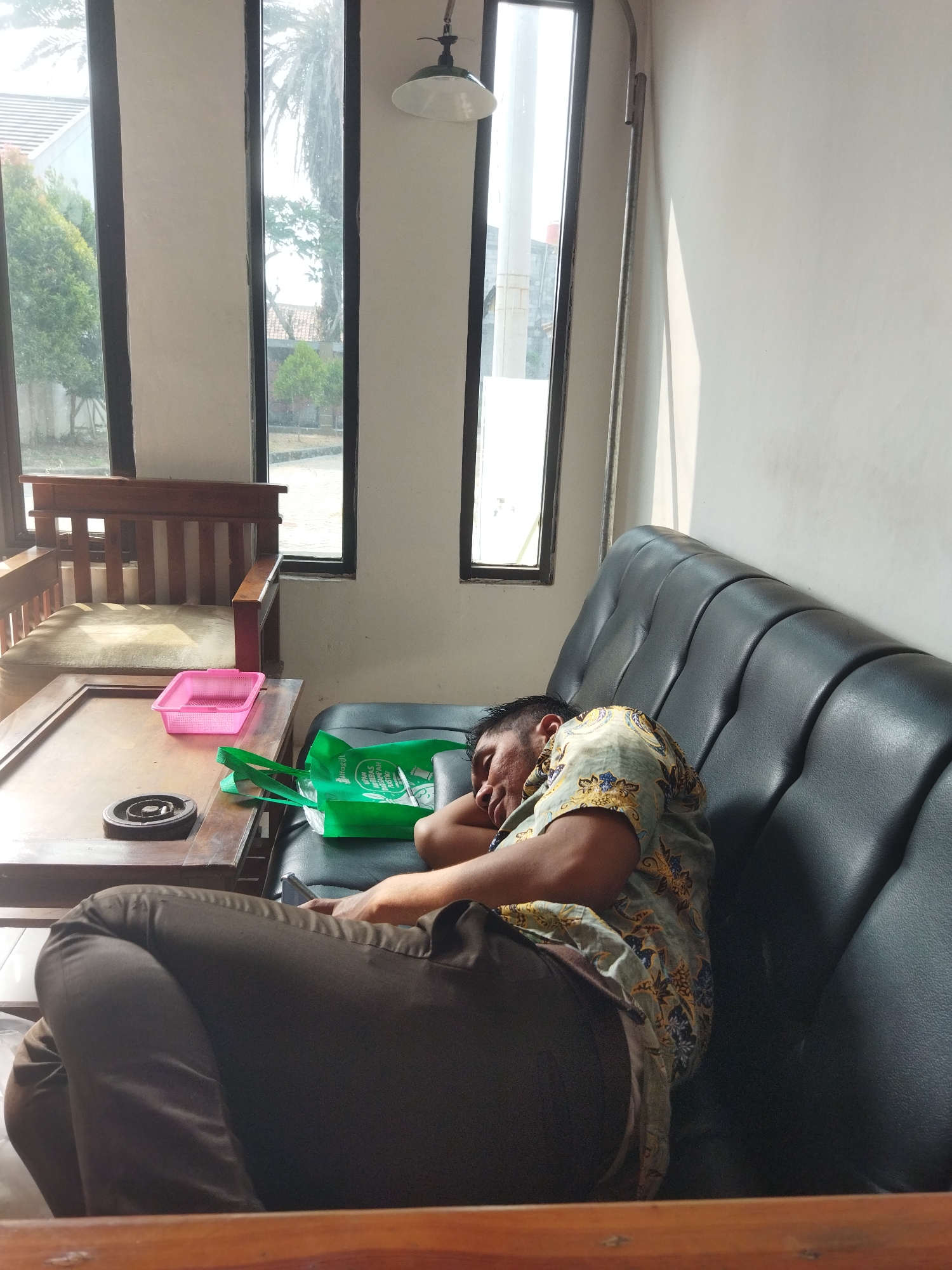 A man is asleep on a black leather couch in what appears to be an indoor waiting area or lounge. He is wearing a short-sleeved patterned shirt and dark pants. To his left, on a wooden coffee table, are a pink plastic container and a green reusable shopping bag. An ashtray is also visible on the table. Behind the couch, a large window with multiple panes looks out onto an outdoor scene with trees and buildings. The room is illuminated by natural light streaming in from the window, casting shadows on the wall. The overall atmosphere is one of quiet repose. The location is Suka Sari, Indonesia.