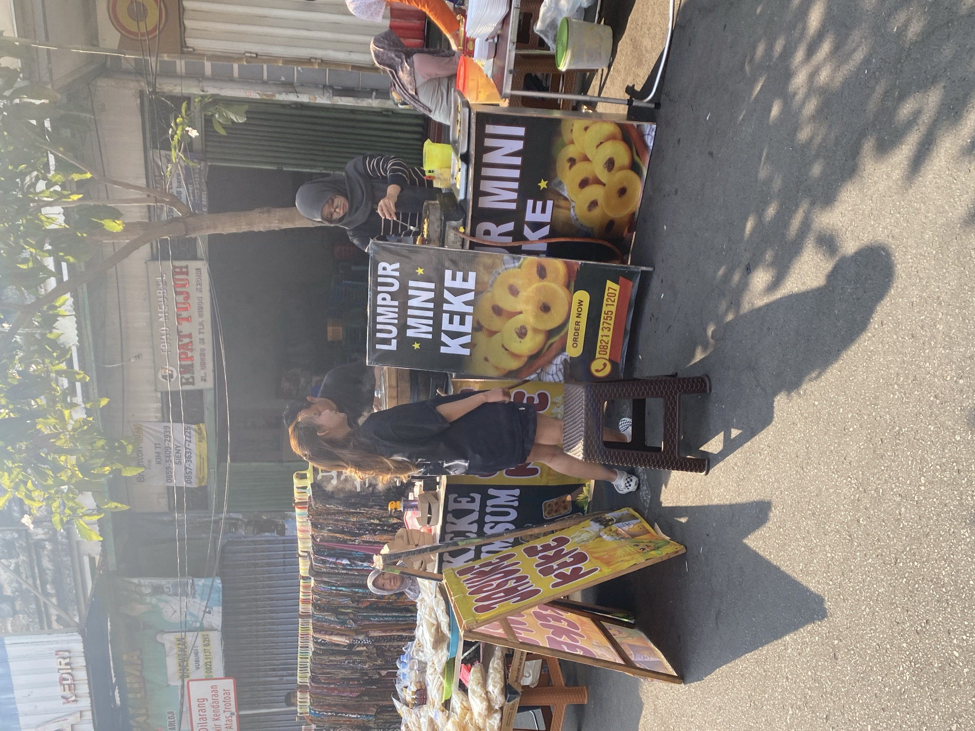 This image depicts a street market scene in Kediri City, Indonesia, likely on a sunny day given the shadows. In the foreground, a young woman with long brown hair, wearing a black t-shirt and shorts, is walking past a food stall. To her left, a stall displays various bags of snacks. Adjacent to this, a colorful A-frame sign with Indonesian text, "JASUKE KEKEE," is prominently featured. Further to the right, a more formal stall has a black banner reading "LUMPUR MINI KEKE" with an image of small yellow pastries. Behind this banner, another similar banner is visible. A woman wearing a headscarf is operating this stall, cooking food on a portable stove. Another woman, also wearing a headscarf, is seen in the background at a different stall, partially obscured. The market is set up along a paved street, with buildings and signage indicating a commercial area. Several other stalls and tables are visible in the background, laden with goods. The overall impression is that of a bustling local market.